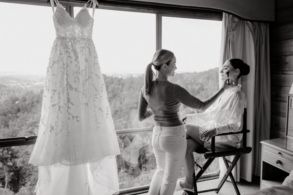 A wedding dress hangs by a window while preparations take place in a bridal suite with scenic views.