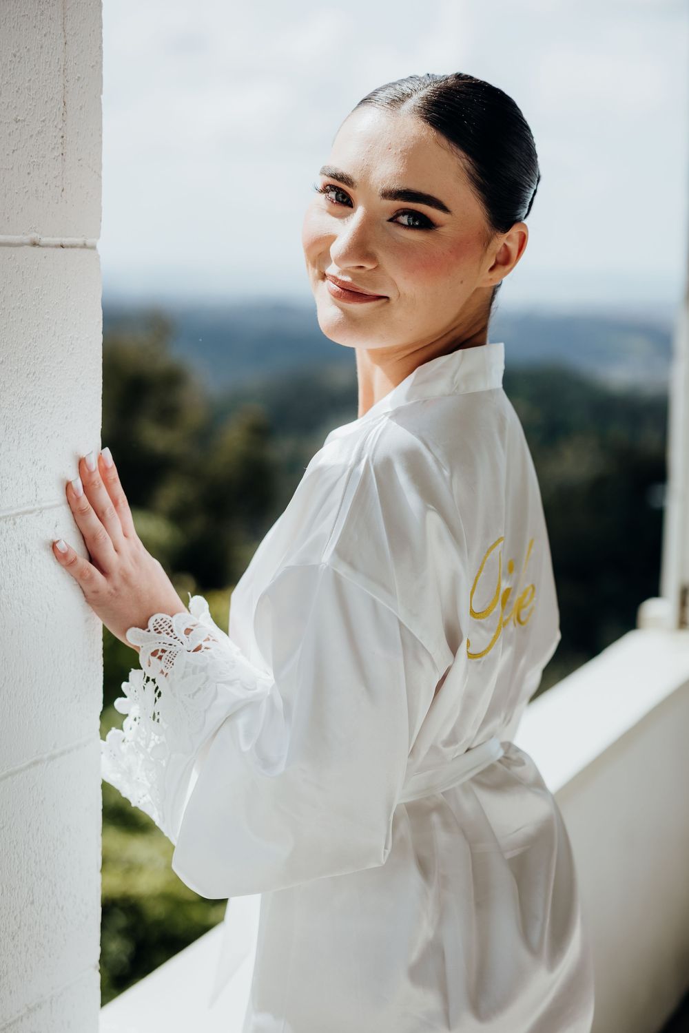 A bride in a white silk robe poses on a balcony with a scenic mountain backdrop.