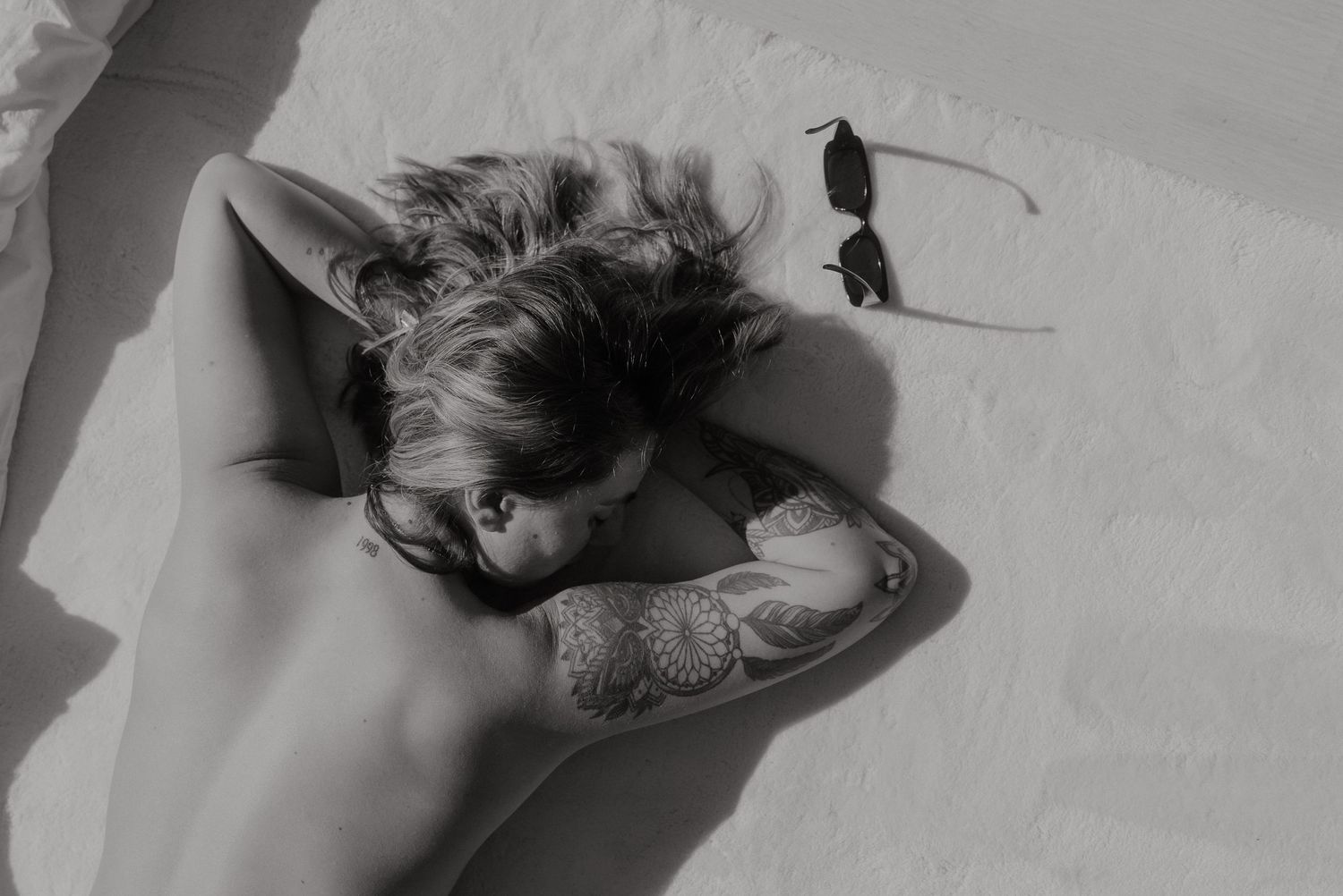 Black and white artistic portrait of person with tattoos resting against wall.