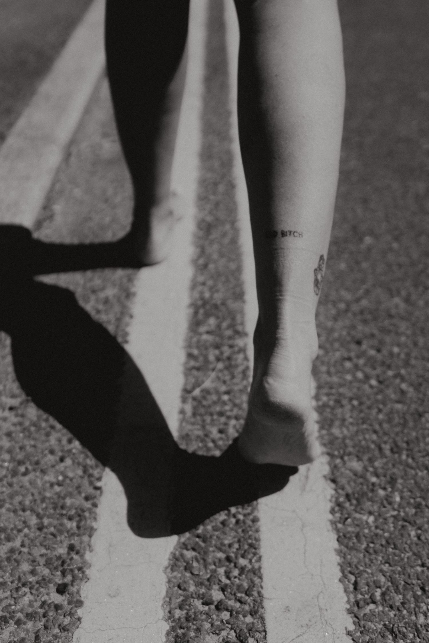 Black and white photo of legs and shadow on pavement with small tattoo visible.
