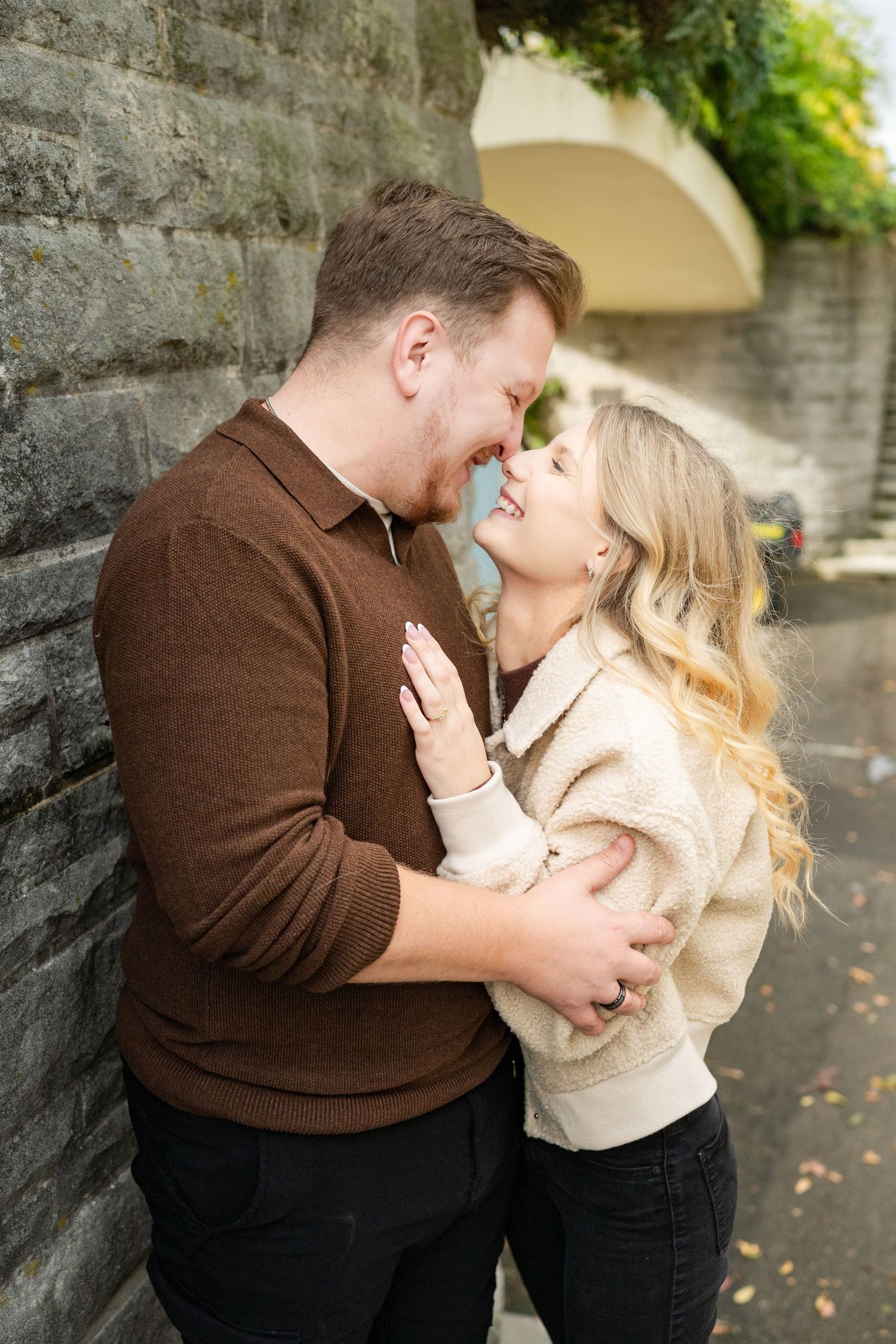 A romantic couple in autumn shares tender moments against a stone wall while wearing brown and cream colored outfits.