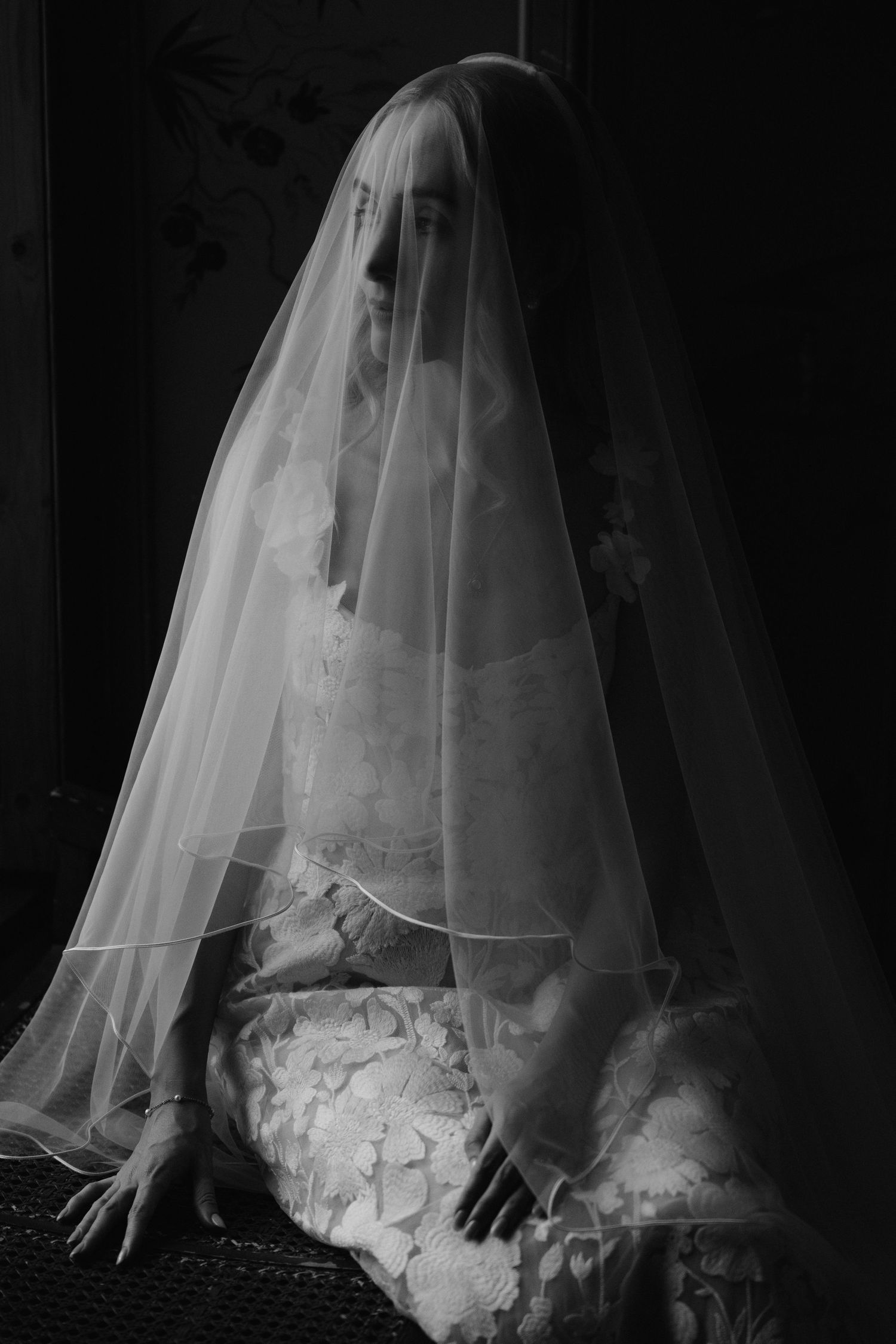 A figure in a flowing white wedding dress and veil sits mysteriously in dramatic black and white lighting.