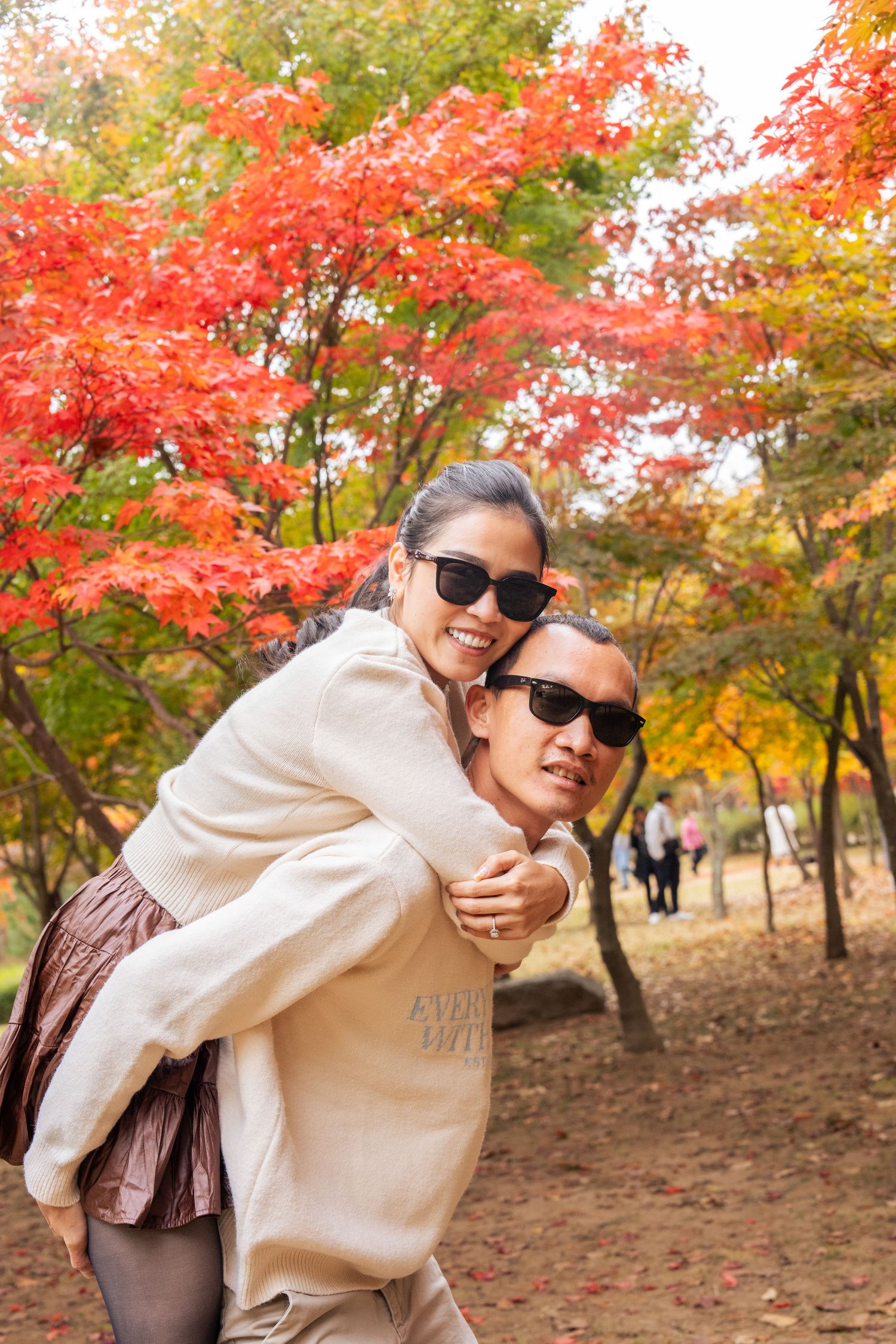 Person in sunglasses and beige sweater poses in front of vibrant autumn maple trees with red foliage in a park setting.
