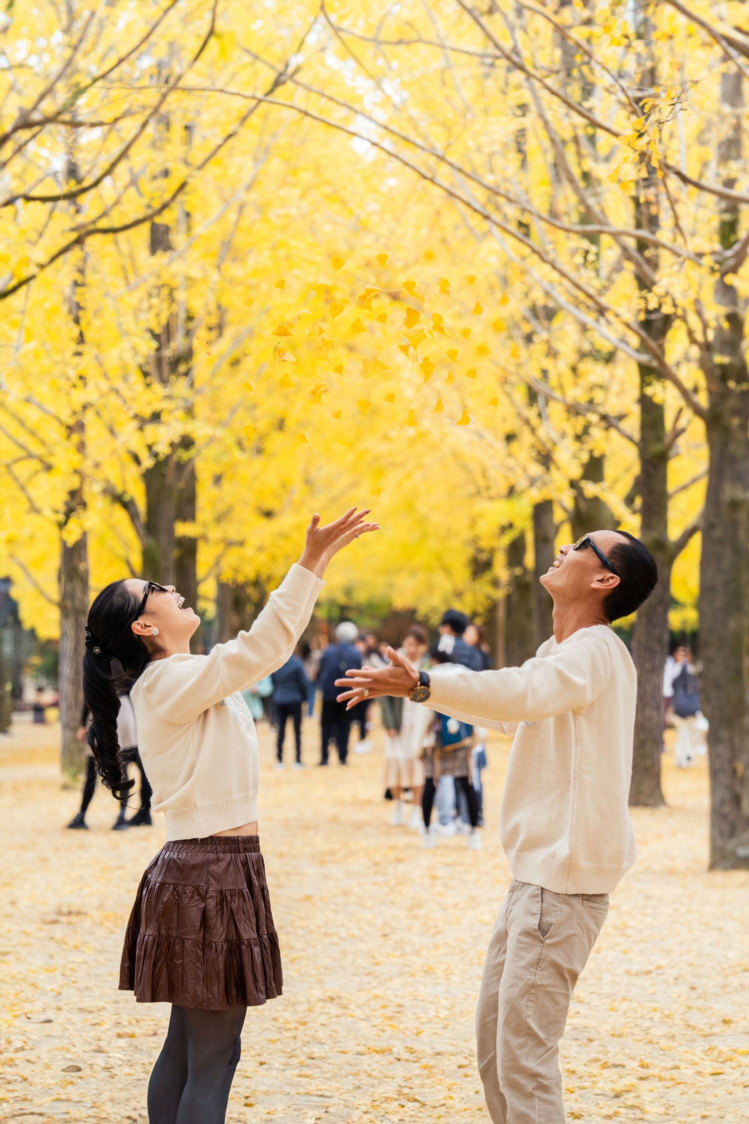 People in white and brown outfits play with falling yellow autumn leaves in a park lined with golden ginkgo trees.