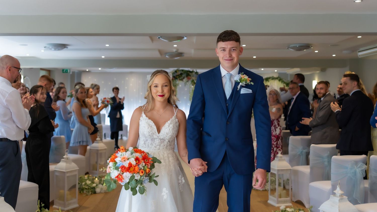 Wedding ceremony guests seated in white chairs at an elegant indoor venue with floral decorations and chandeliers.