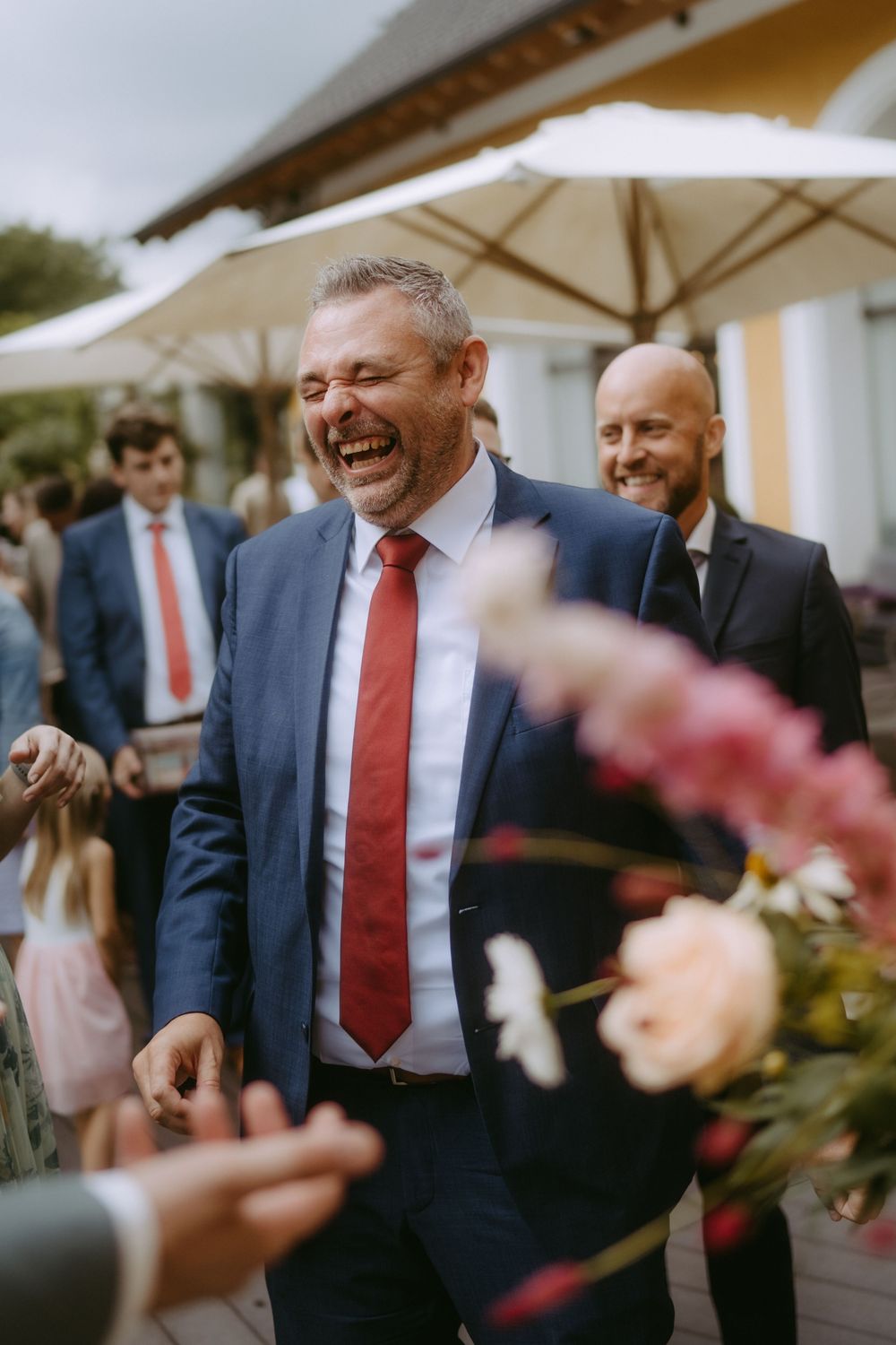 Wedding guests in suits and red ties laughing during outdoor reception festivities.
