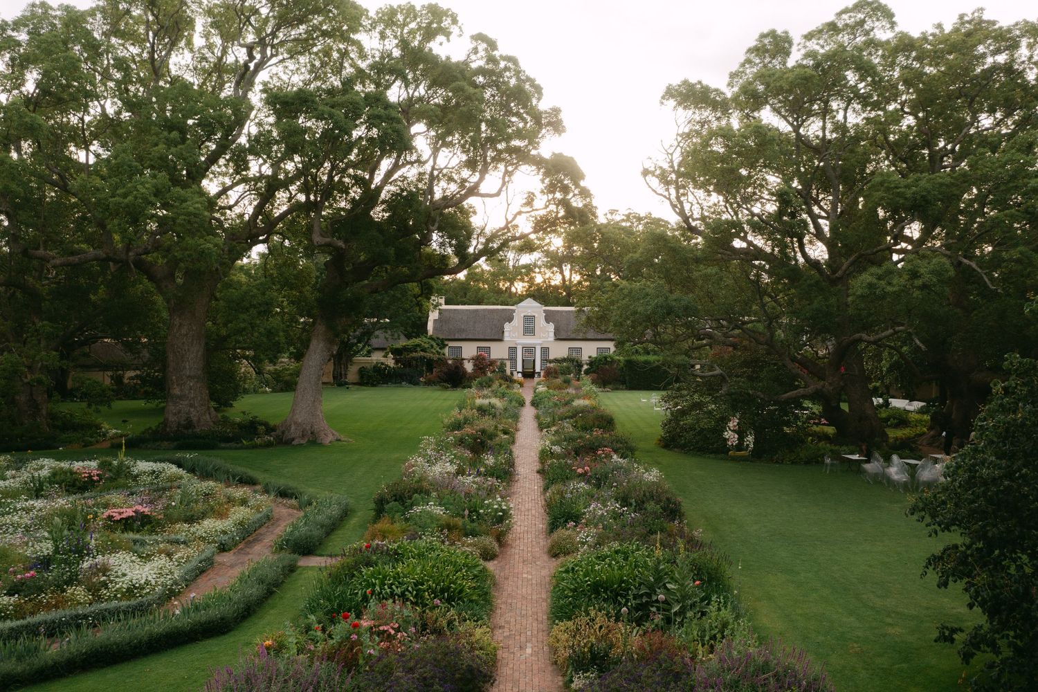 A scenic garden pathway leads to a stately white mansion surrounded by lush green trees and manicured landscaping.