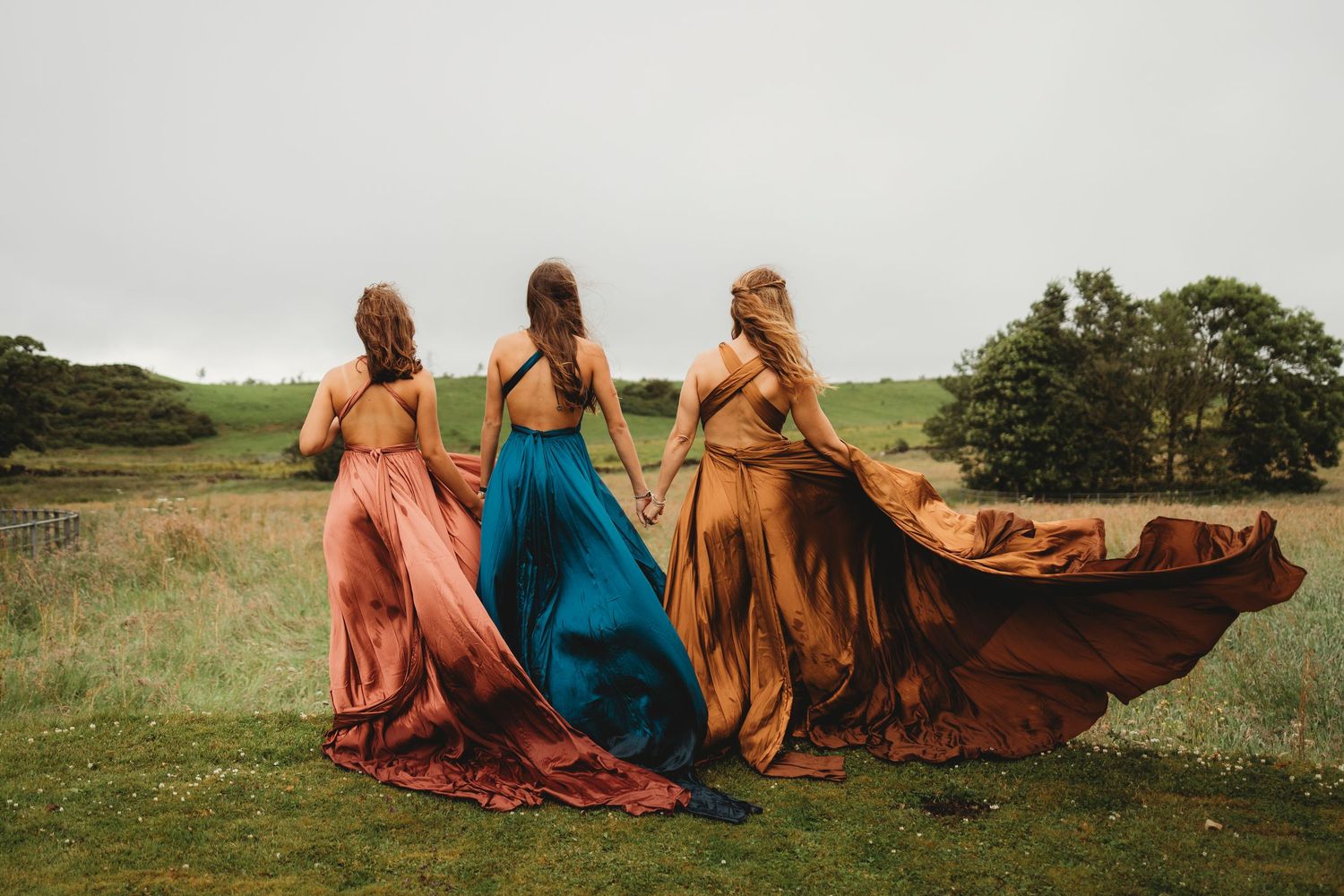 Four people in flowing dresses sit together on a grassy hillside during golden hour.