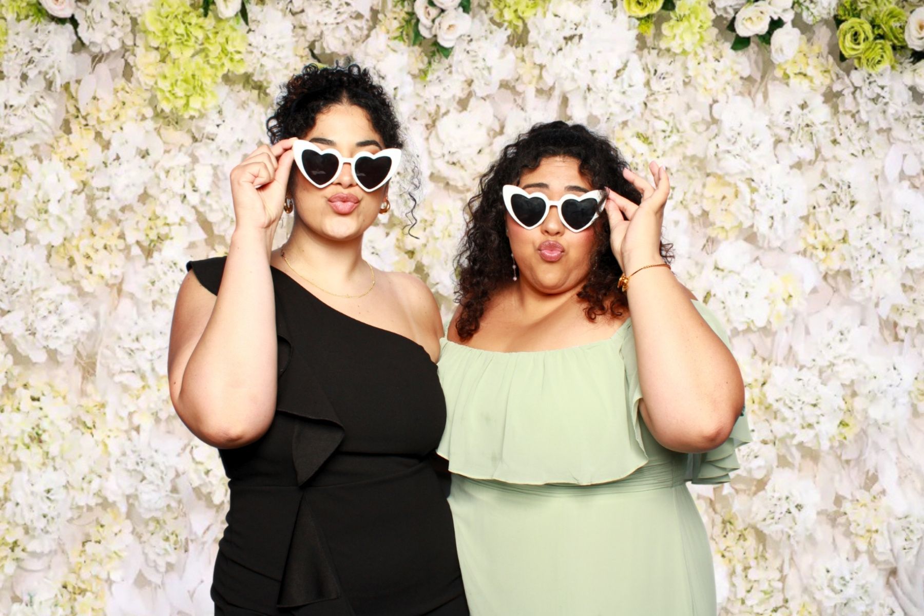 Two people in sunglasses pose playfully in front of a floral wall backdrop at a photo booth.