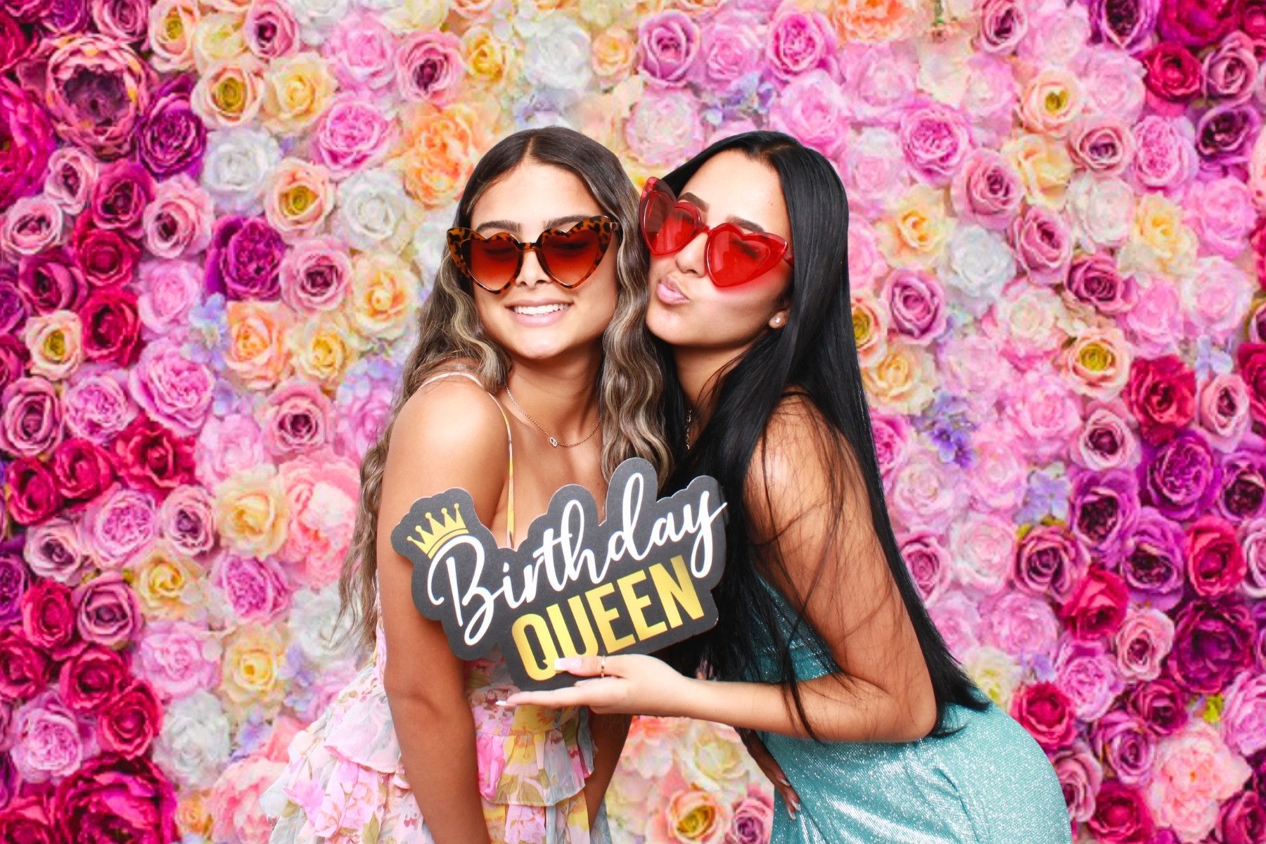 Two friends pose together against a vibrant pink floral wall backdrop at a birthday celebration.