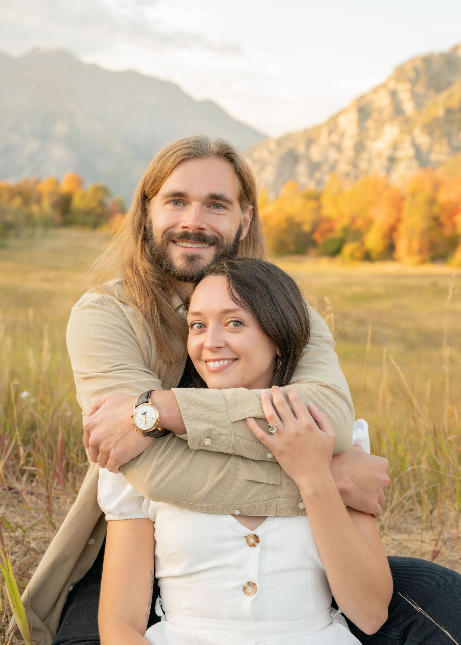 A couple embraces in a golden field at sunset with mountains in the background during an outdoor photo session.