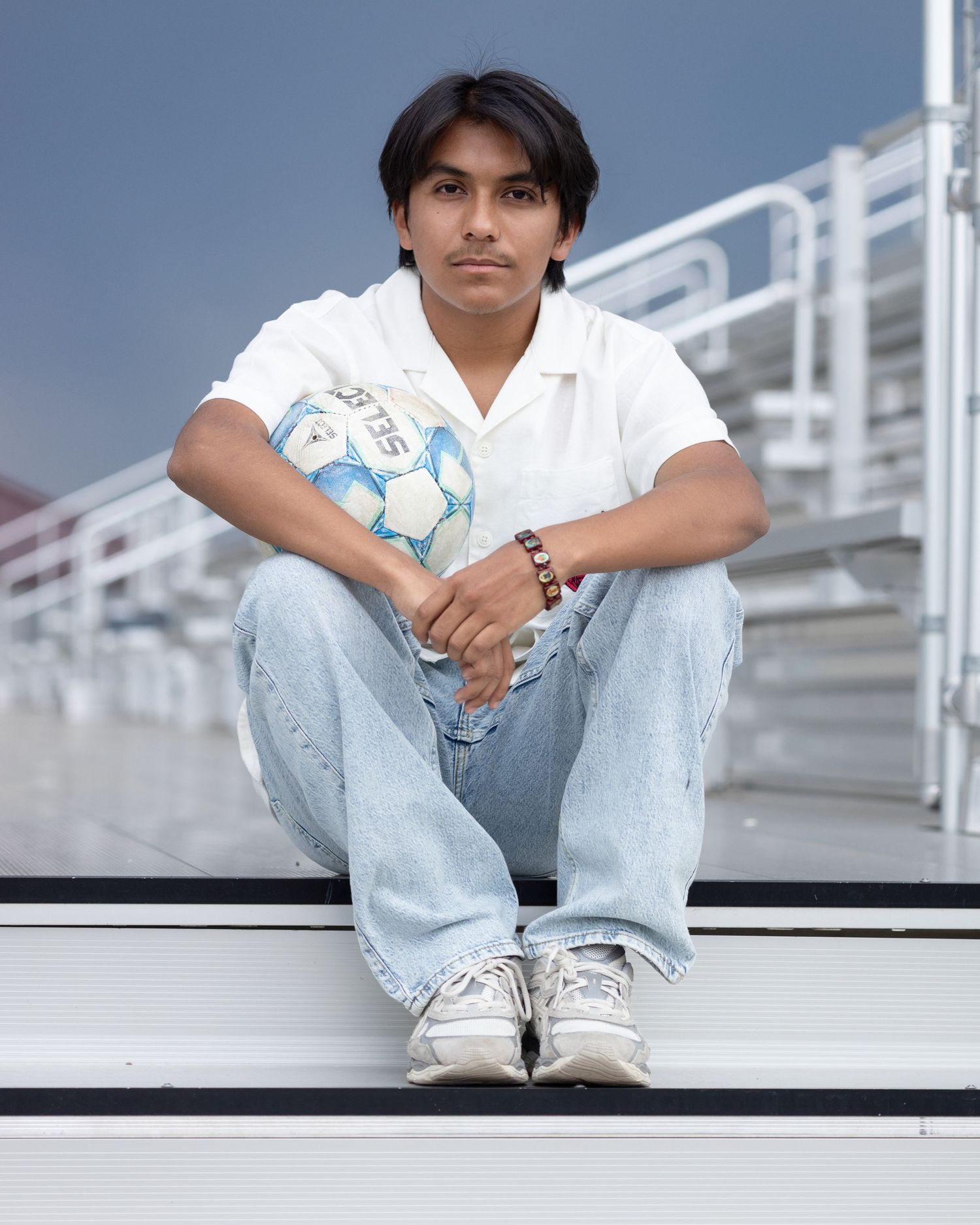 A person sits on bleacher steps holding a soccer ball while wearing light jeans, a white polo shirt and tennis shoes.