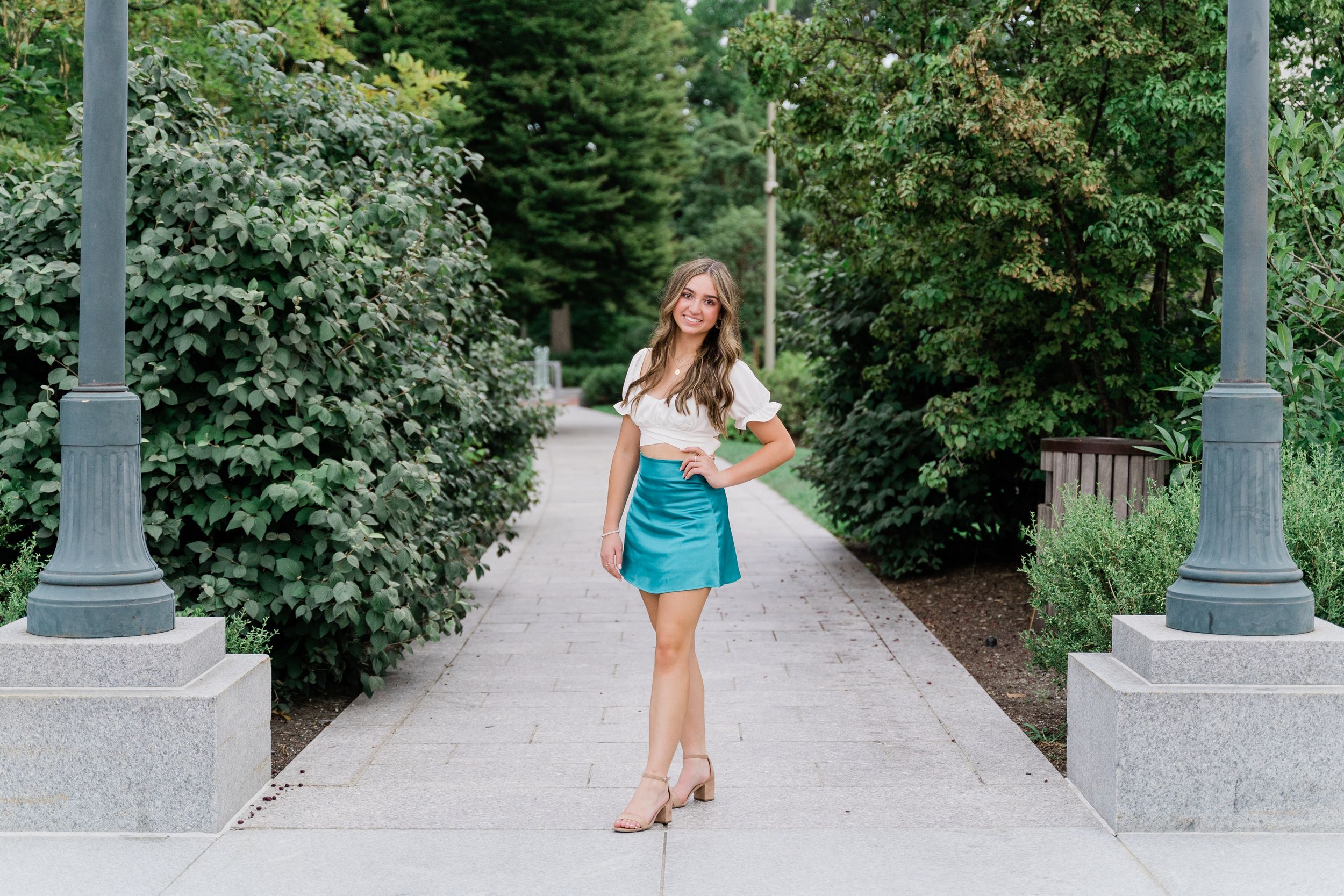 A person in a teal dress walks along a stone path bordered by lush greenery and decorative lamp posts.