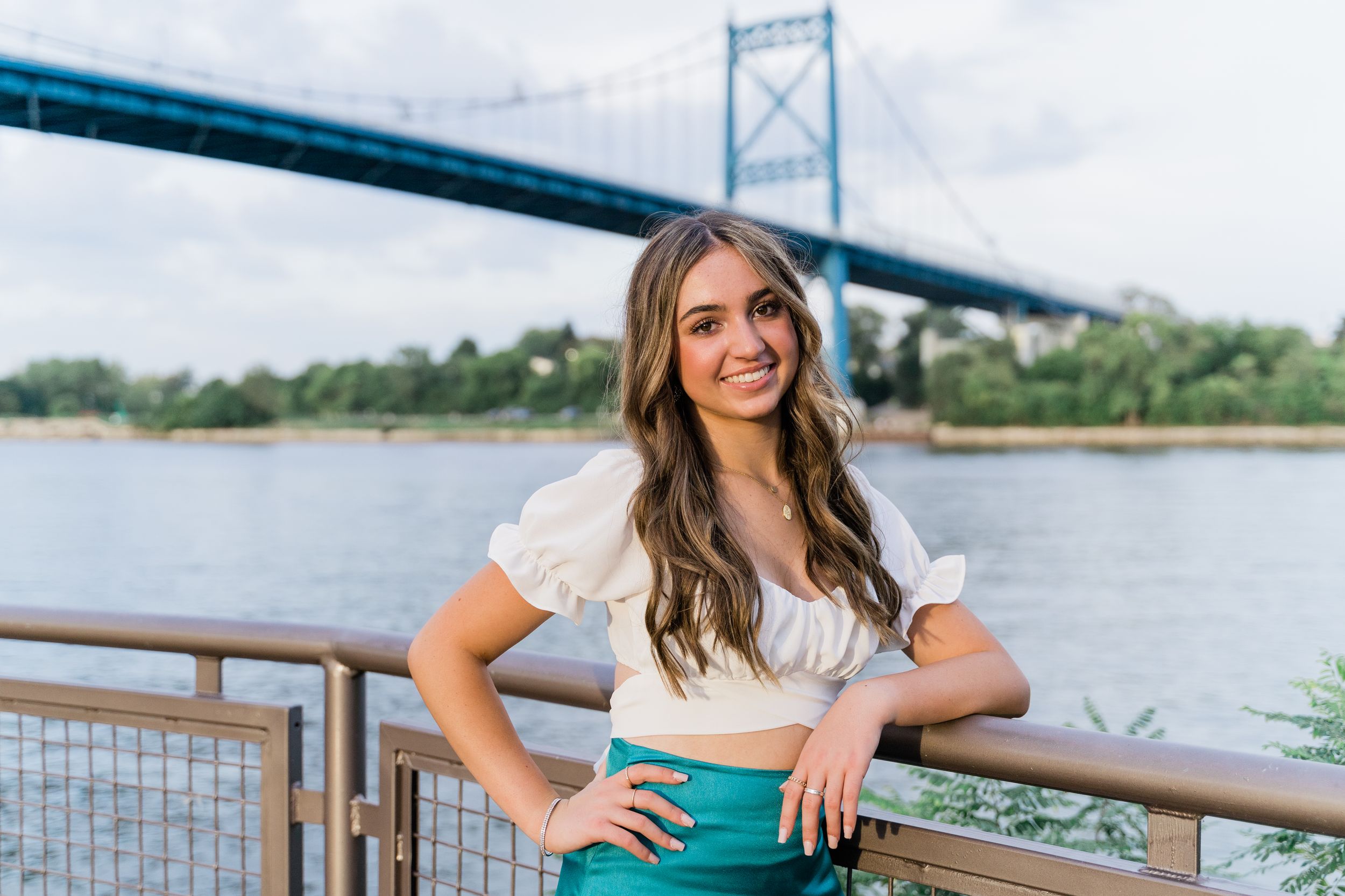 A person in a teal dress poses for outdoor photos near a bridge and waterfront on a sunny day.