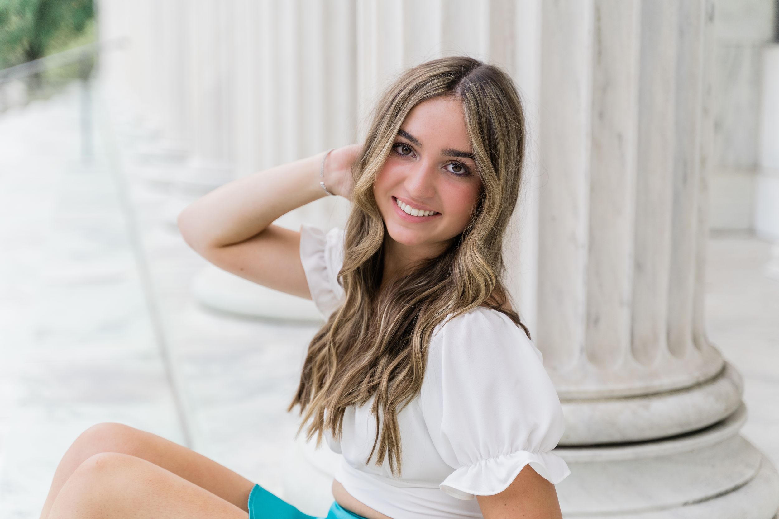 A person in a white top sits near classic white columns with long wavy hair and a bright smile.
