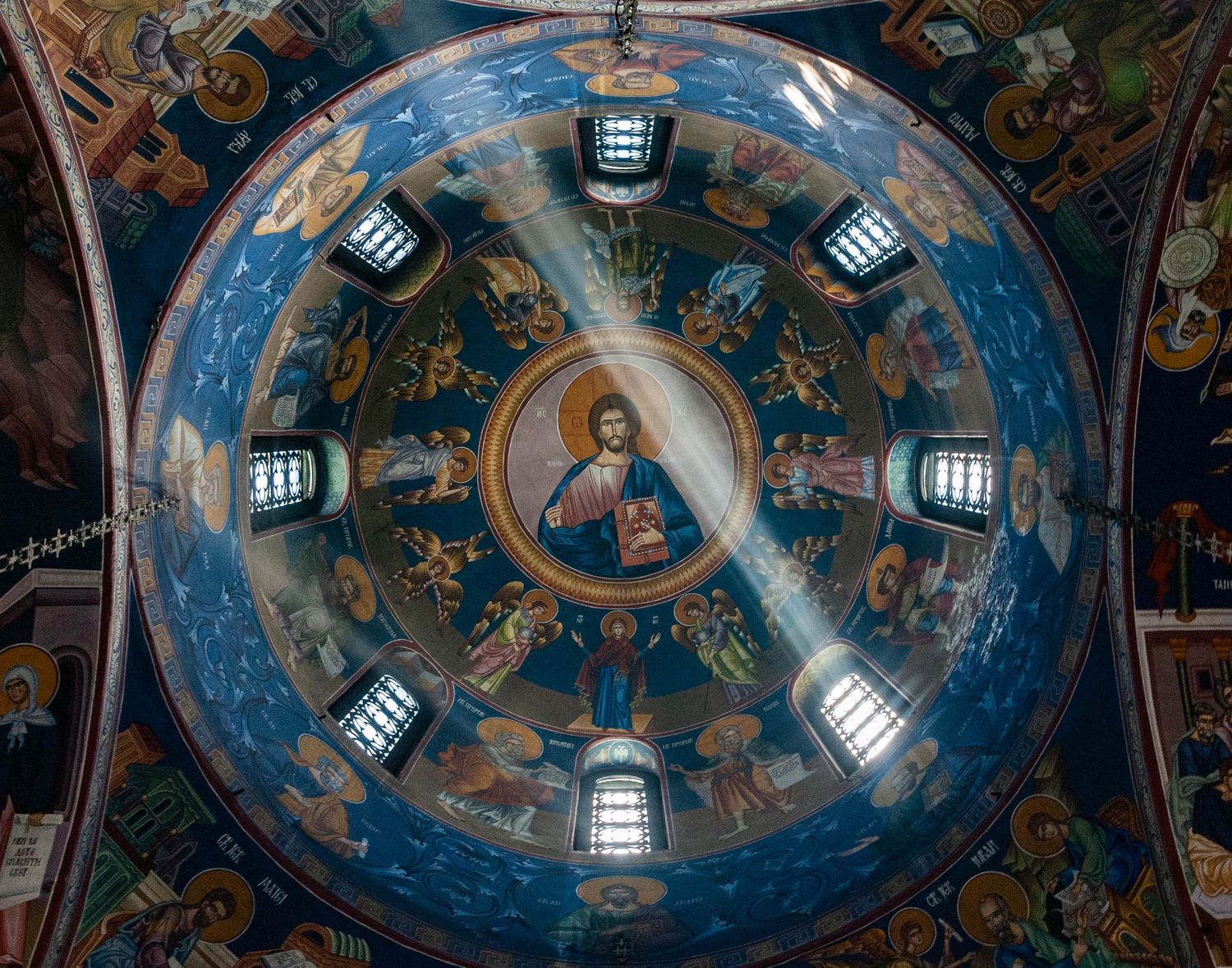 Ornate religious frescoes and iconography adorn the domed ceiling of an Orthodox Christian church with natural light streaming in.