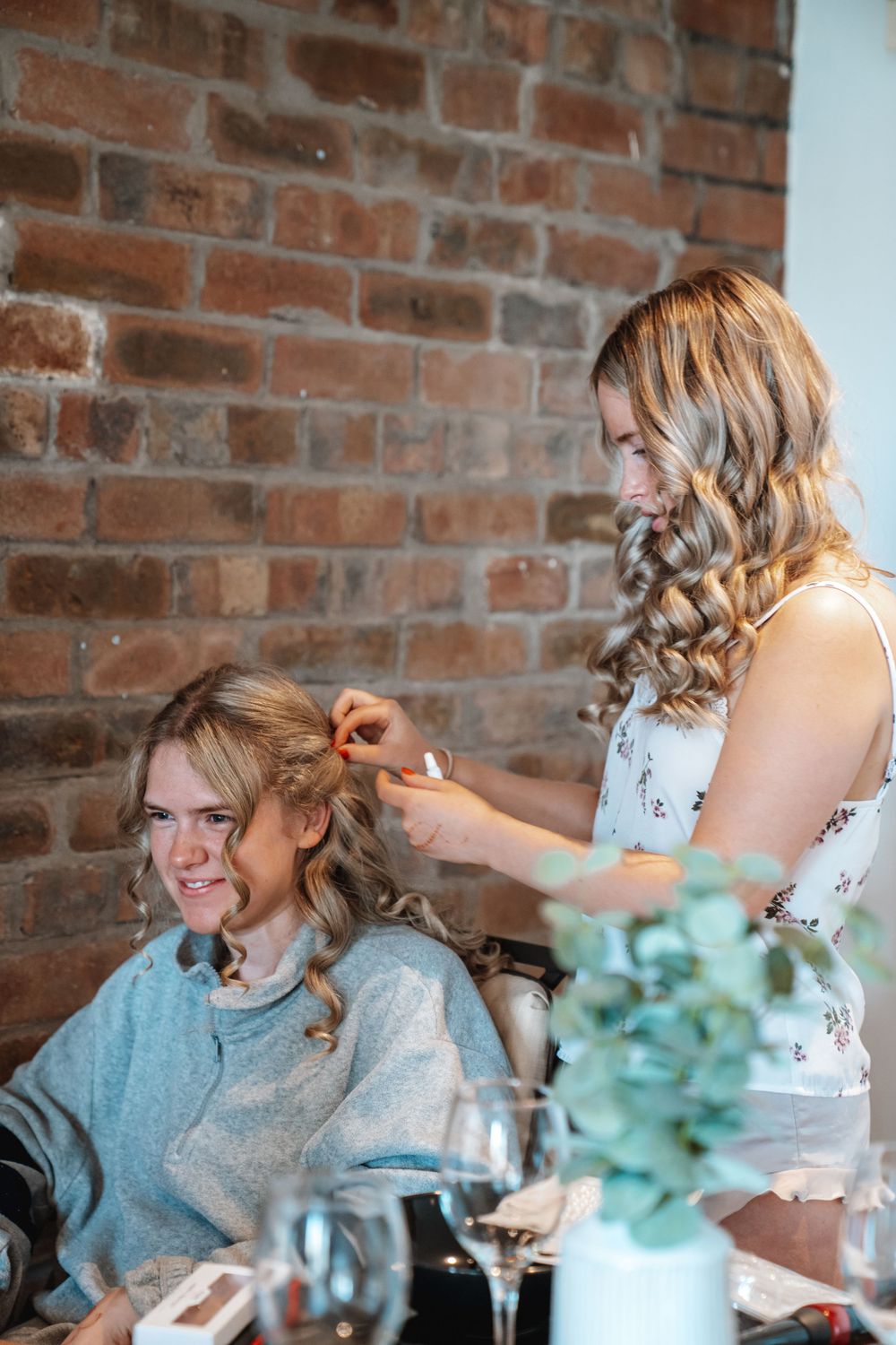 Hairstylist with curly blonde hair styling bridesmaids hair at table with eucalyptus decor and brick wall backdrop.