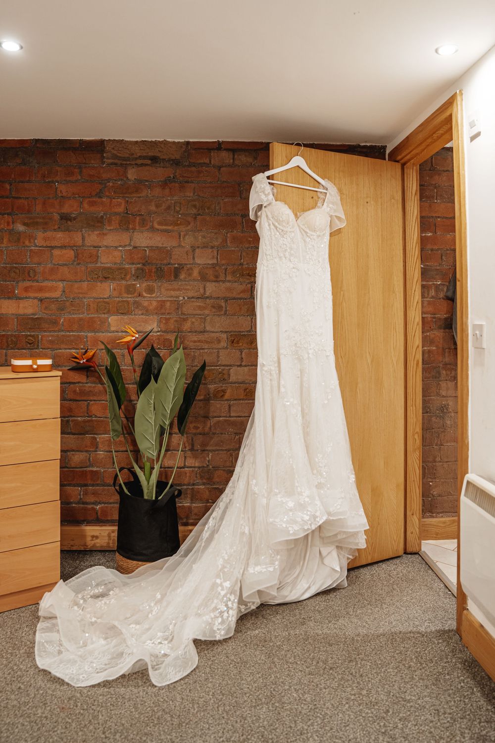 Elegant lace wedding dress with long train hanging on wooden door beside brick wall and tropical plant.