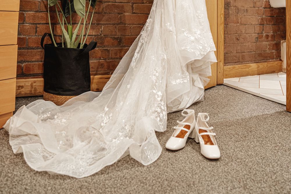 White lace wedding dress train draped on floor beside white Mary Jane bridal shoes near brick wall.