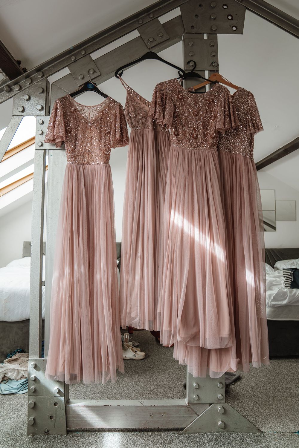 Three dusty rose bridesmaid dresses with sequin bodices and tulle skirts hanging on industrial rack in sunlit room.