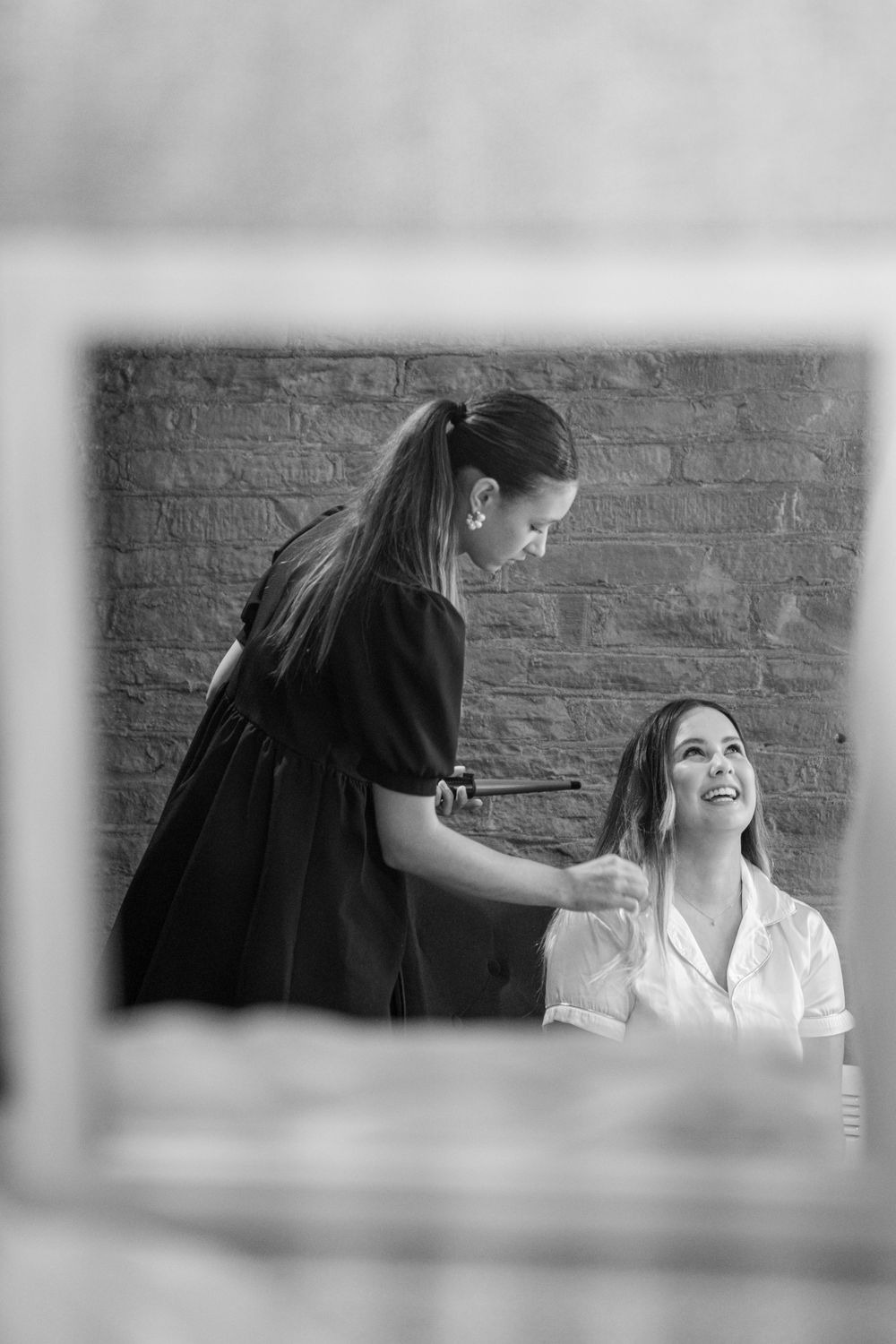 Black and white photo of a hairstylist curling a smiling womans hair in front of a brick wall.