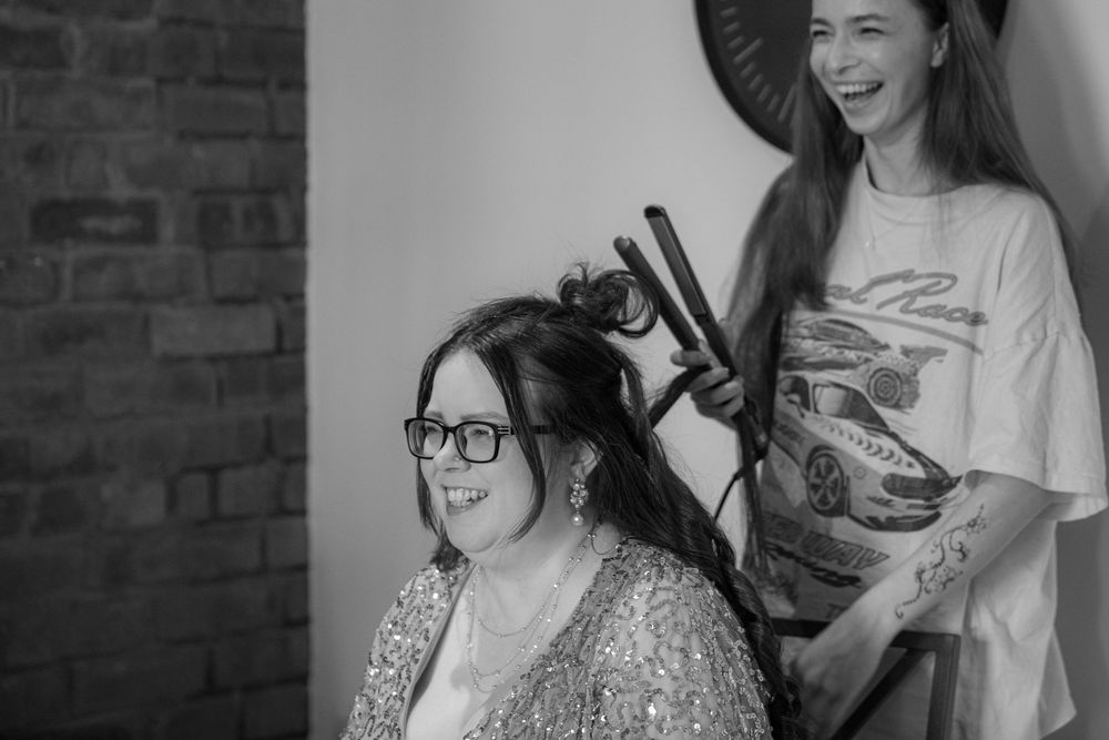Black and white photo of a woman laughing while getting her hair styled with a curling iron.
