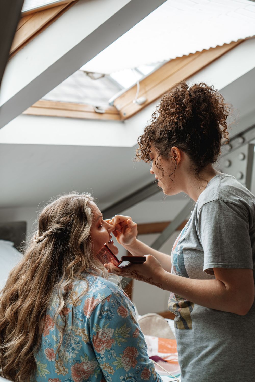 Makeup artist applying eyeshadow to a bride with long wavy hair in a floral robe during wedding preparations.