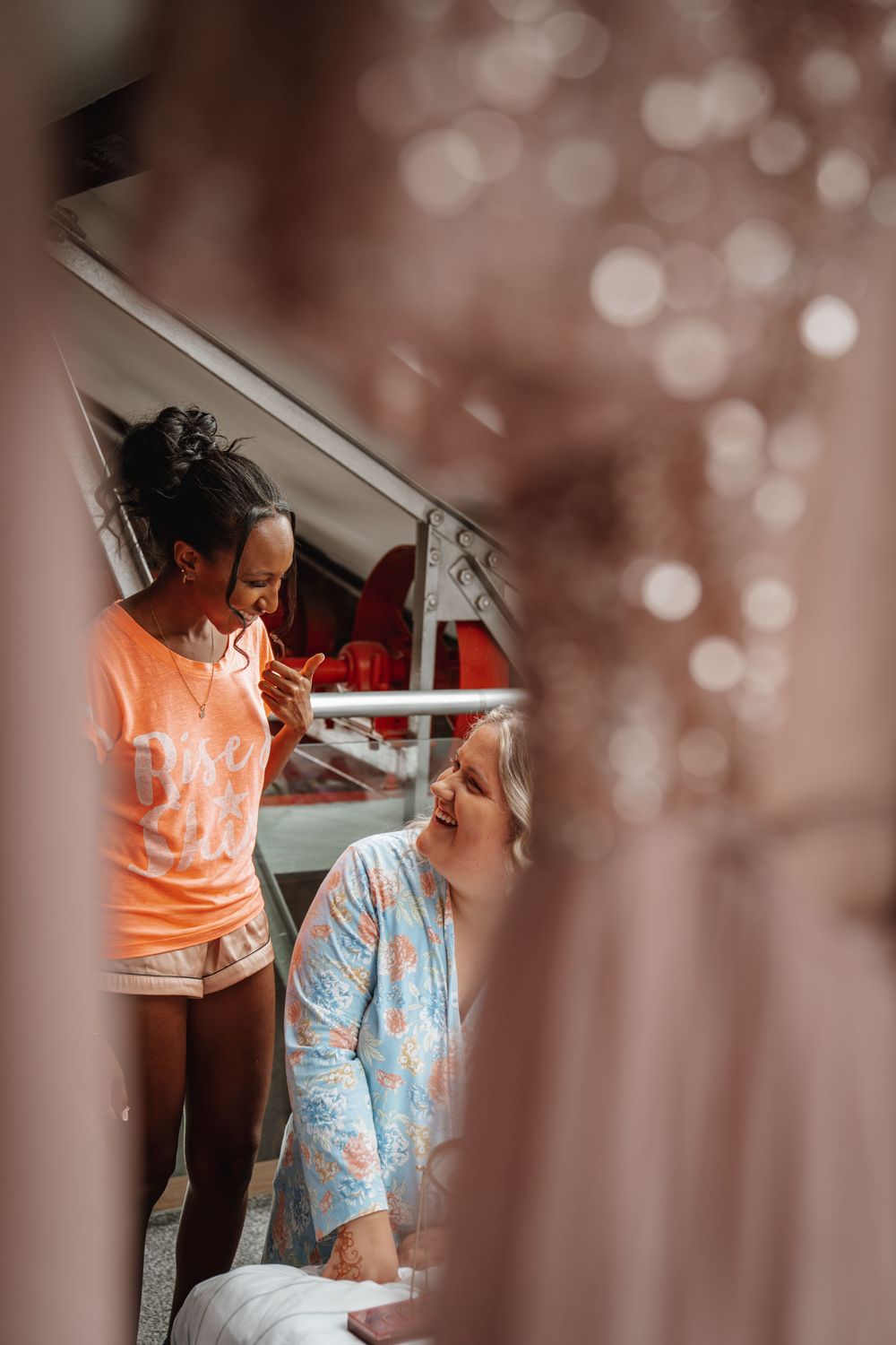 Bridesmaid in orange shirt applying makeup to smiling bride in floral robe, viewed through hanging dresses.