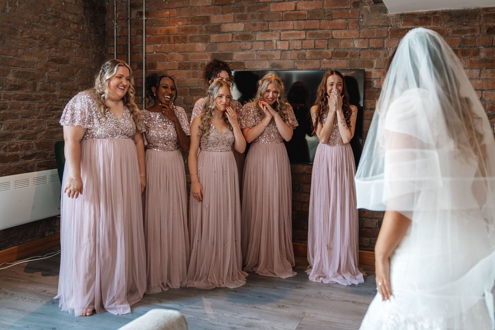 Bridesmaids in blush pink dresses react with emotional surprise as bride in white gown and veil appears.