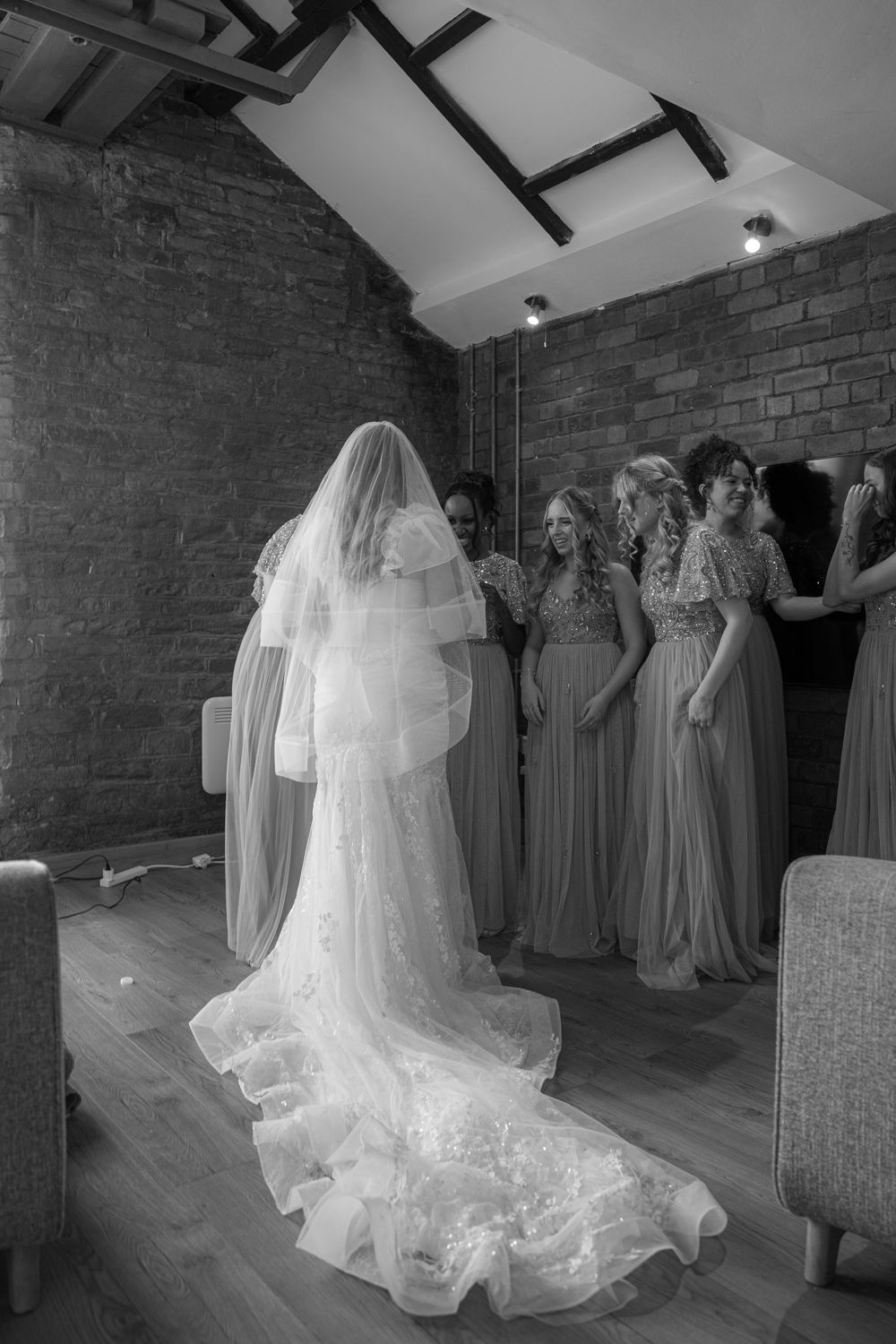 Black and white photo of a bride in a lace gown and veil with bridesmaids in a brick-walled room.
