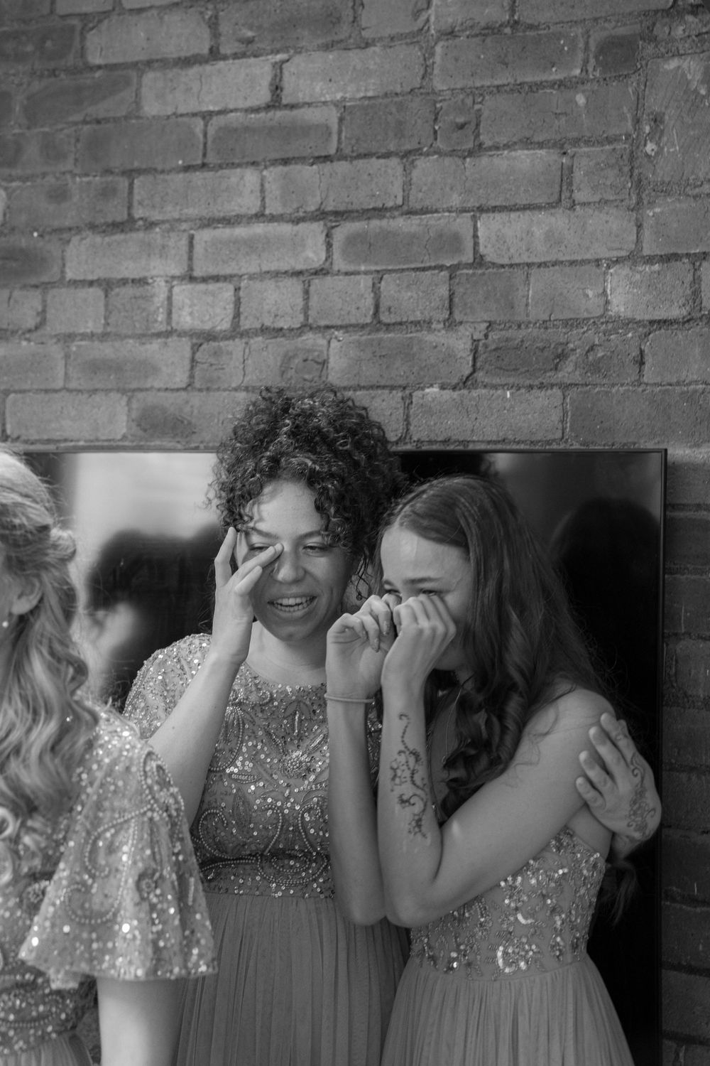 Black and white photo of two emotional bridesmaids wiping tears in front of a brick wall.