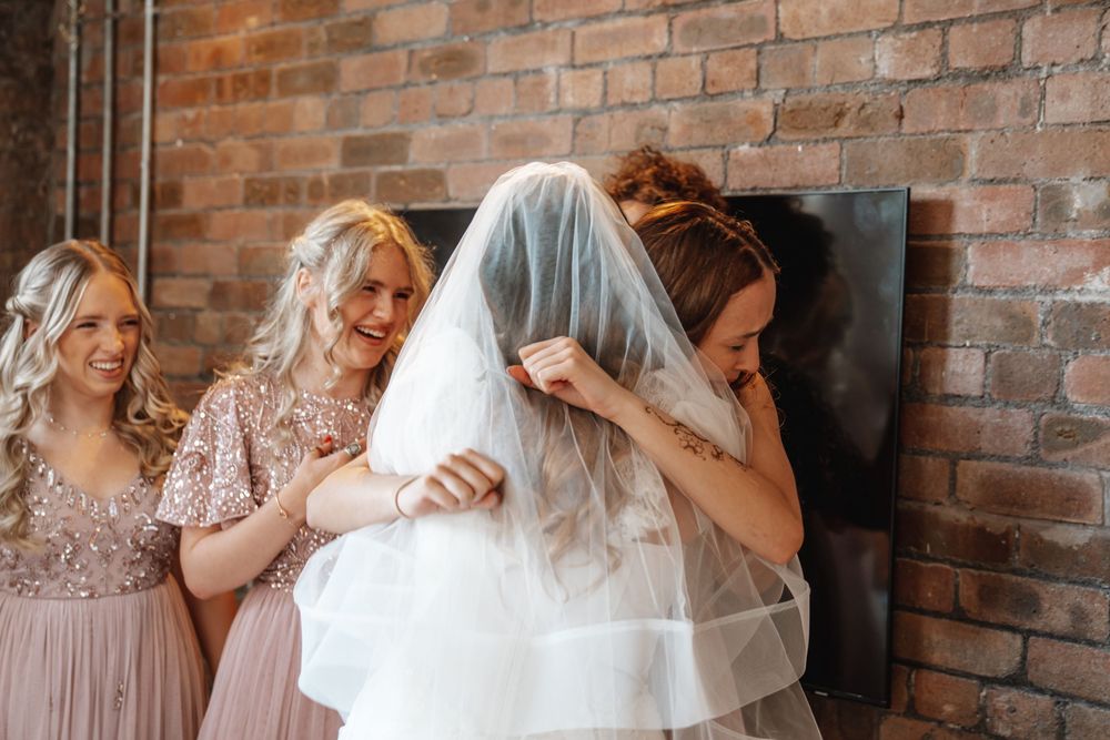Bridesmaids in pink sequin dresses helping bride adjust her white wedding veil against a brick wall.