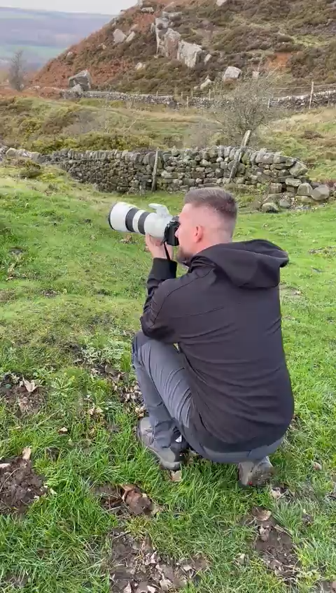 Wildlife photographer lying in grass with telephoto lens capturing nature on mountainous terrain.
