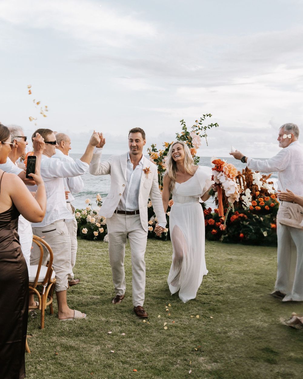 Guests celebrate at an outdoor wedding reception on a grassy hilltop with floral arrangements under cloudy skies.