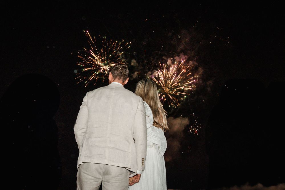 Couple in white clothing embraces while watching fireworks light up the dark night sky.