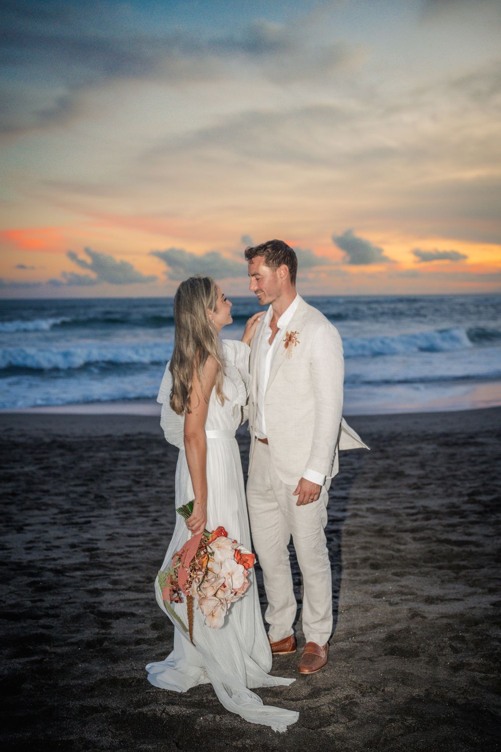A couple in wedding attire stands on a dark beach at sunset with crashing waves and orange sky in the background.
