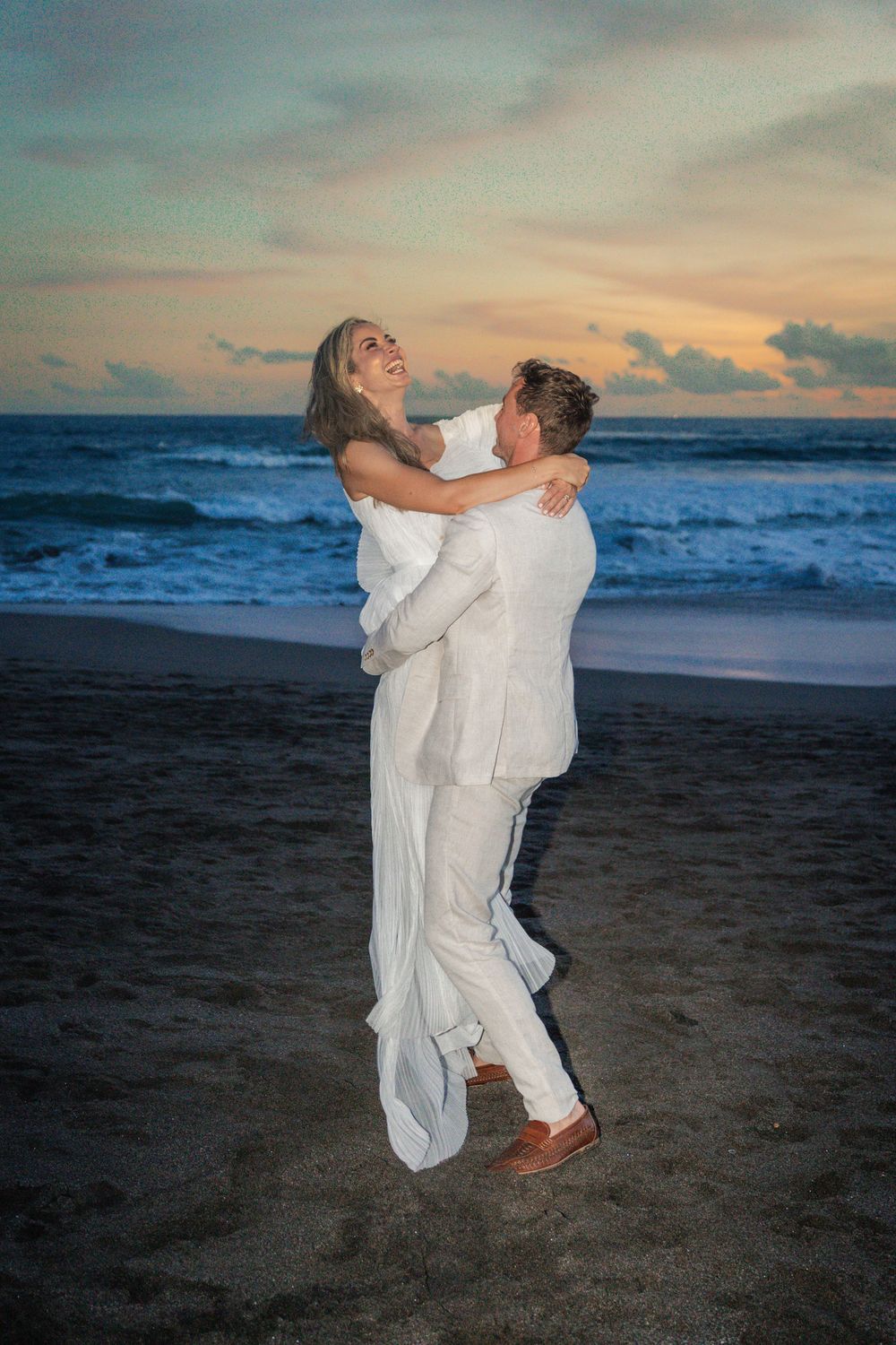 A romantic couple in white clothing embrace on a beach at sunset with ocean waves in the background.