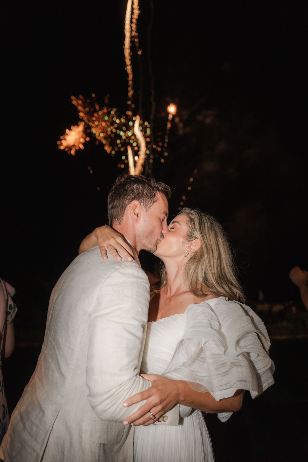 A romantic couple in white attire share a kiss during an evening celebration with twinkling lights in the background.