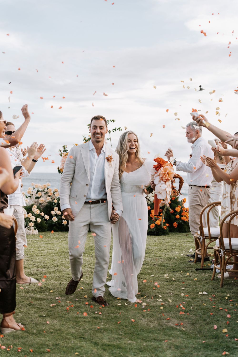 Wedding celebration on a grassy lawn with guests tossing coral-colored flower petals in the air.