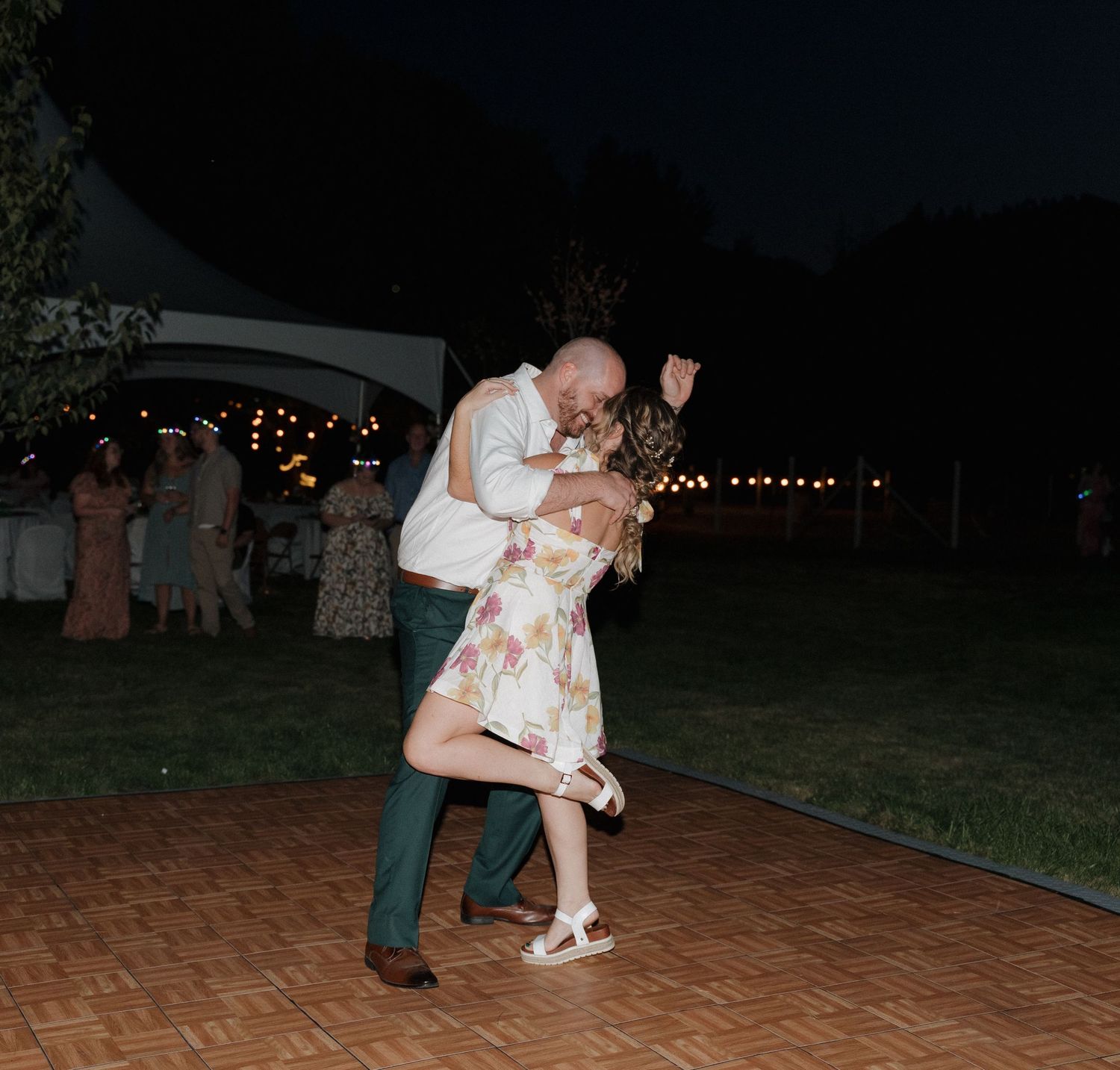 A couple dancing together at an outdoor evening reception on a brick patio with string lights overhead.