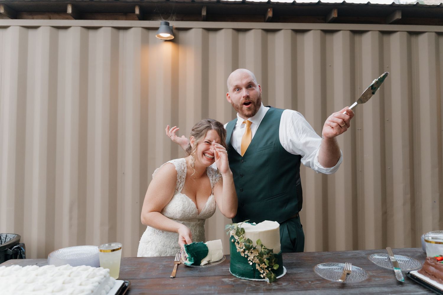 Newlyweds playfully cut their wedding cake together at a reception against a beige wall with overhead lighting.