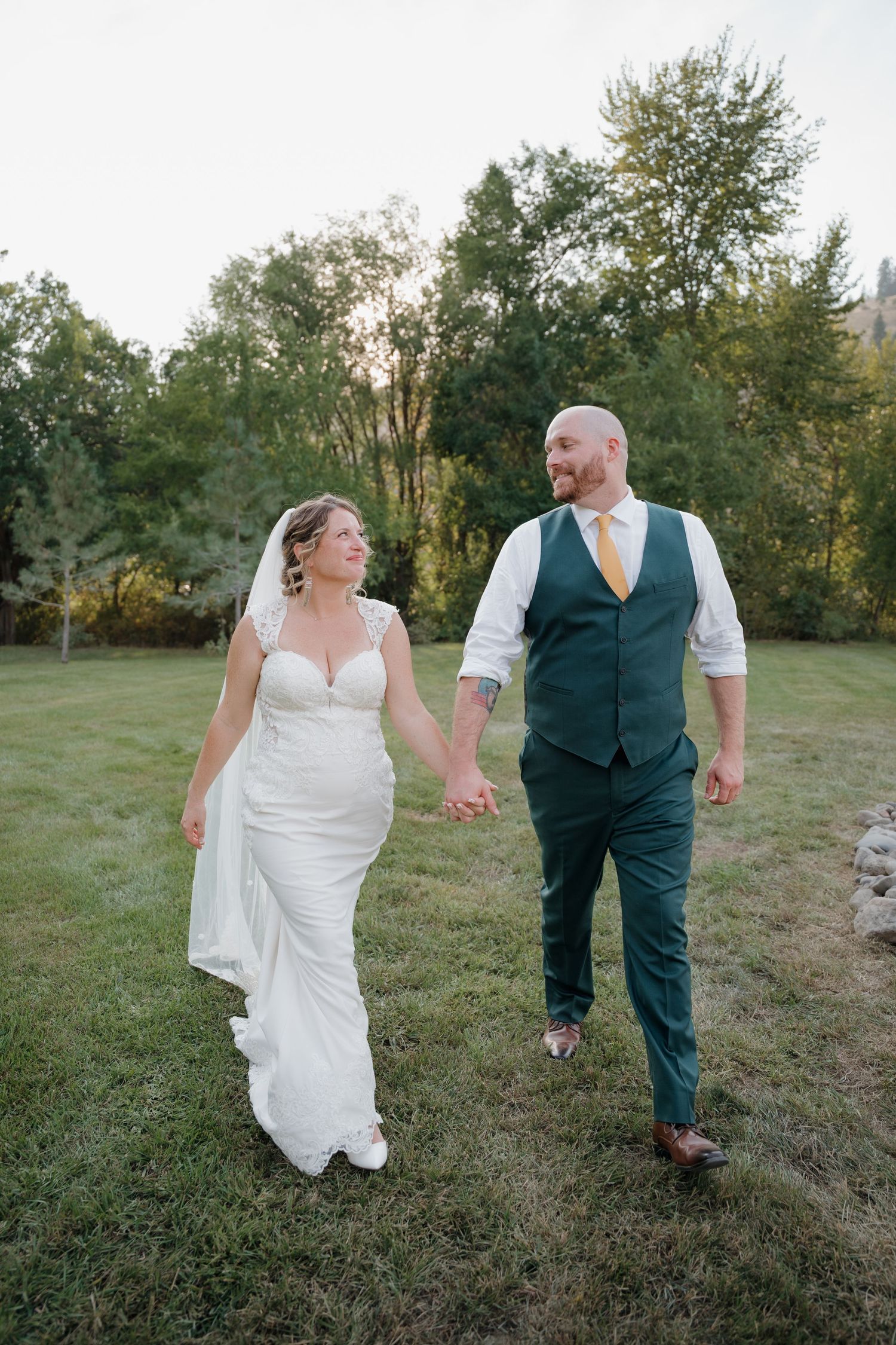 A couple walks hand in hand through a grassy outdoor setting during their wedding celebration at sunset.