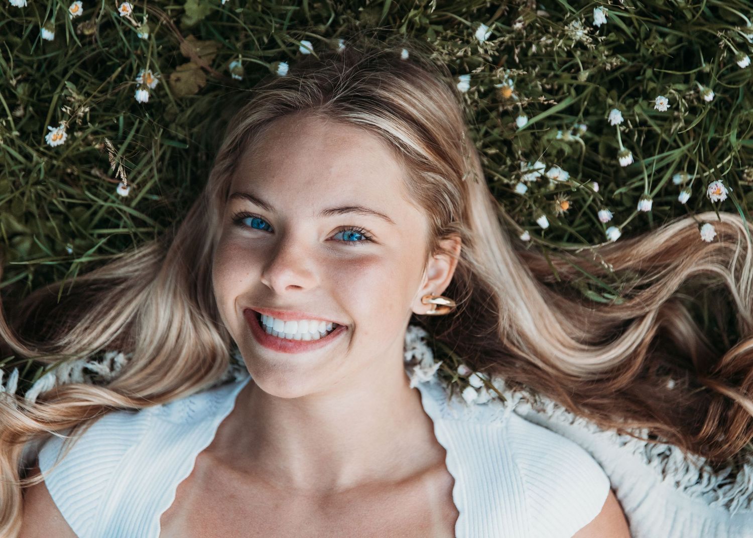 A person lies in white flowers and greenery while their blonde hair fans out around them in a dreamy portrait.
