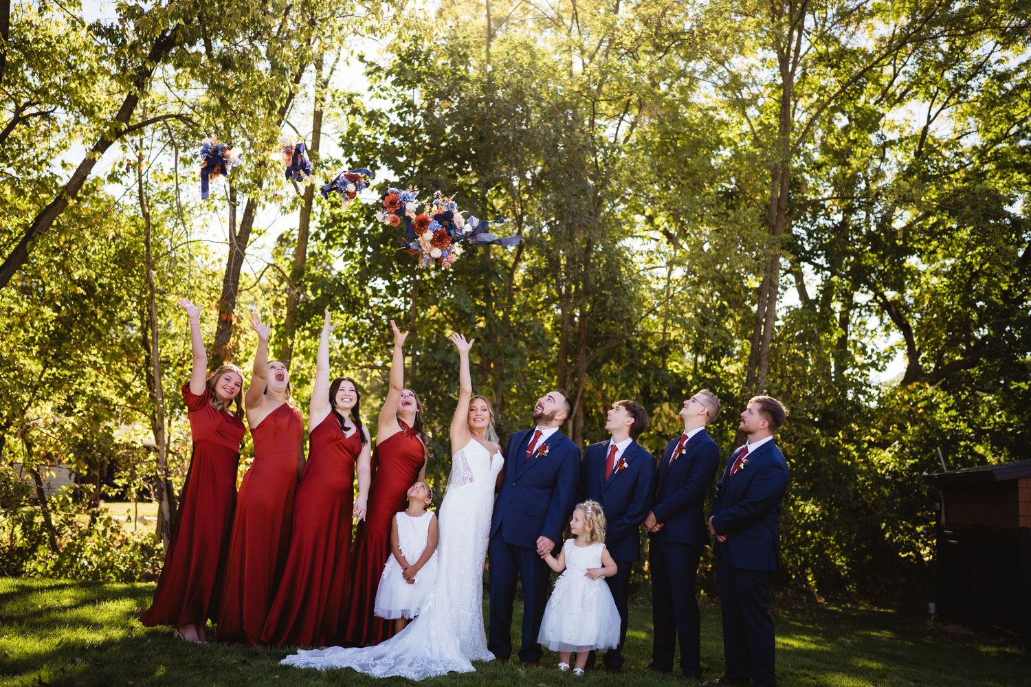 Wedding party poses outdoors in burgundy and navy attire under sunlit trees during autumn ceremony.