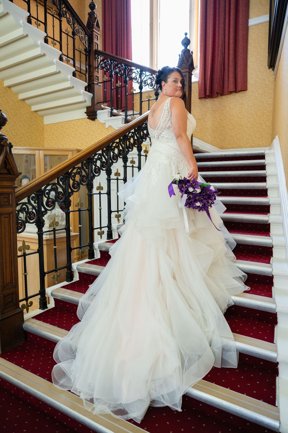Flowing white wedding dress on grand staircase with red carpet and ornate railing.