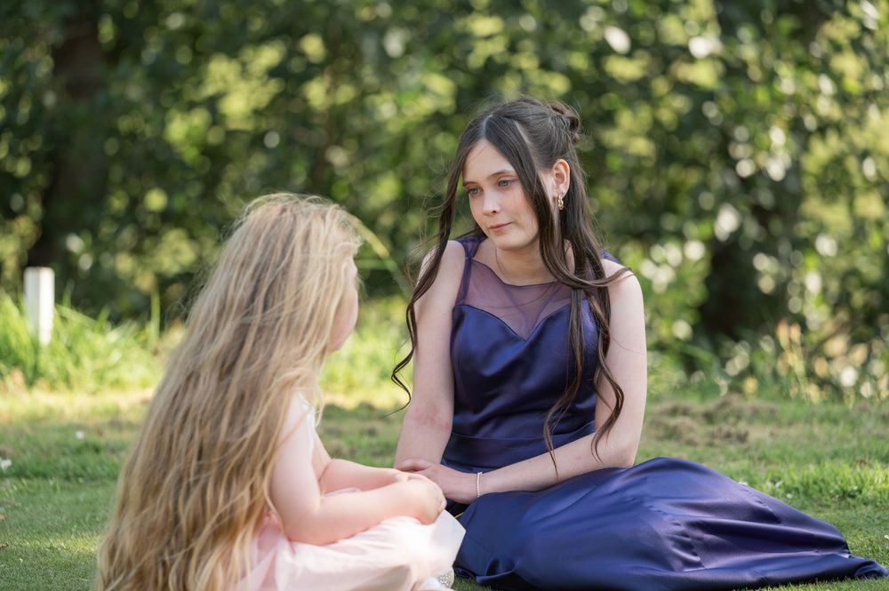 Two people in elegant dresses sit together on grass having an intimate conversation.