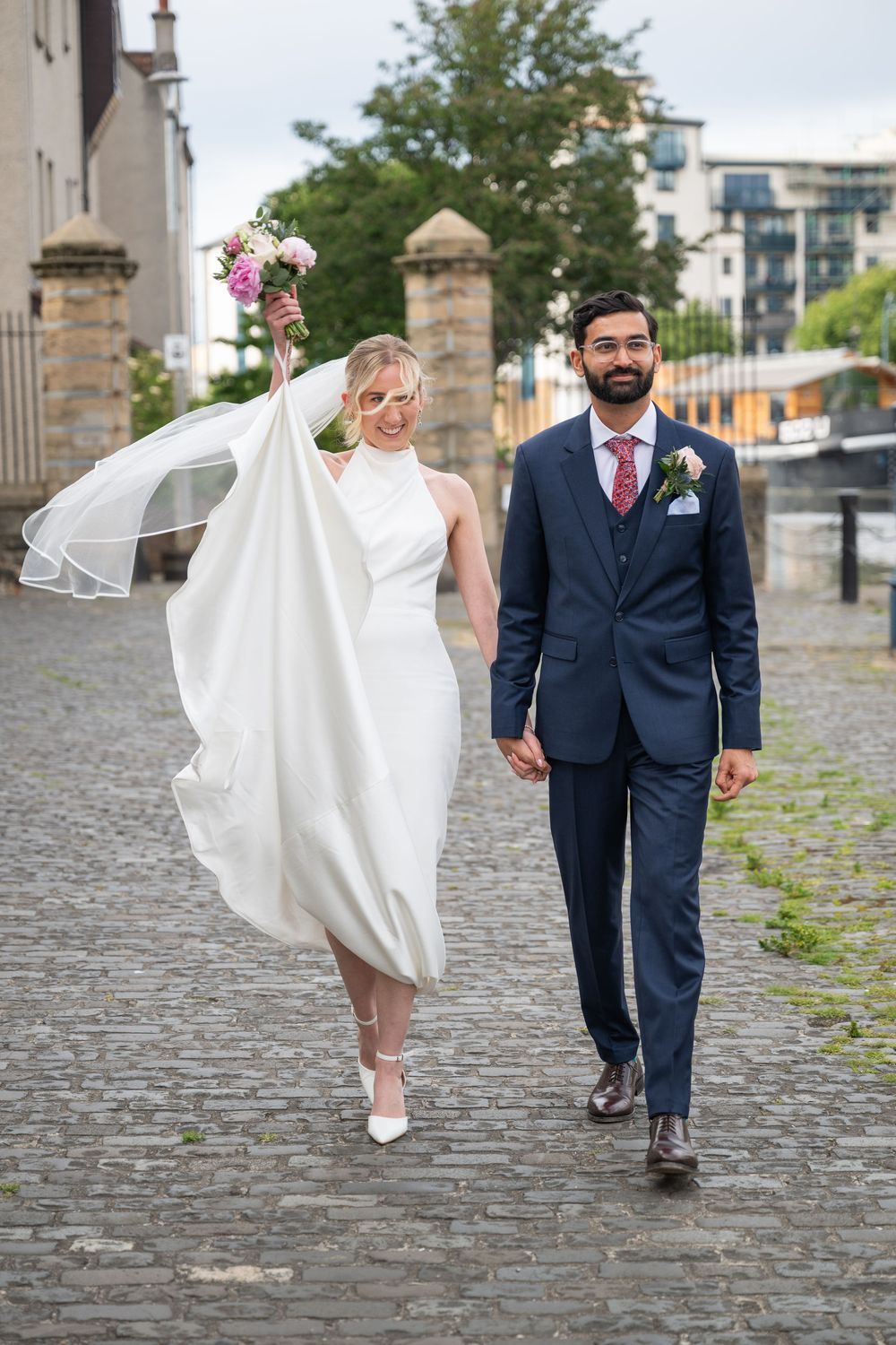 Joyful wedding couple walking on cobblestones with bride's dress and veil flowing in the wind.