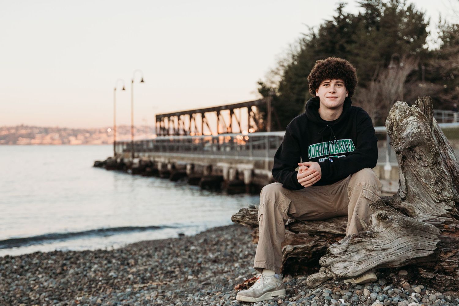 A person sits casually on a rocky beach shoreline during sunset with water and mountains visible in the background.