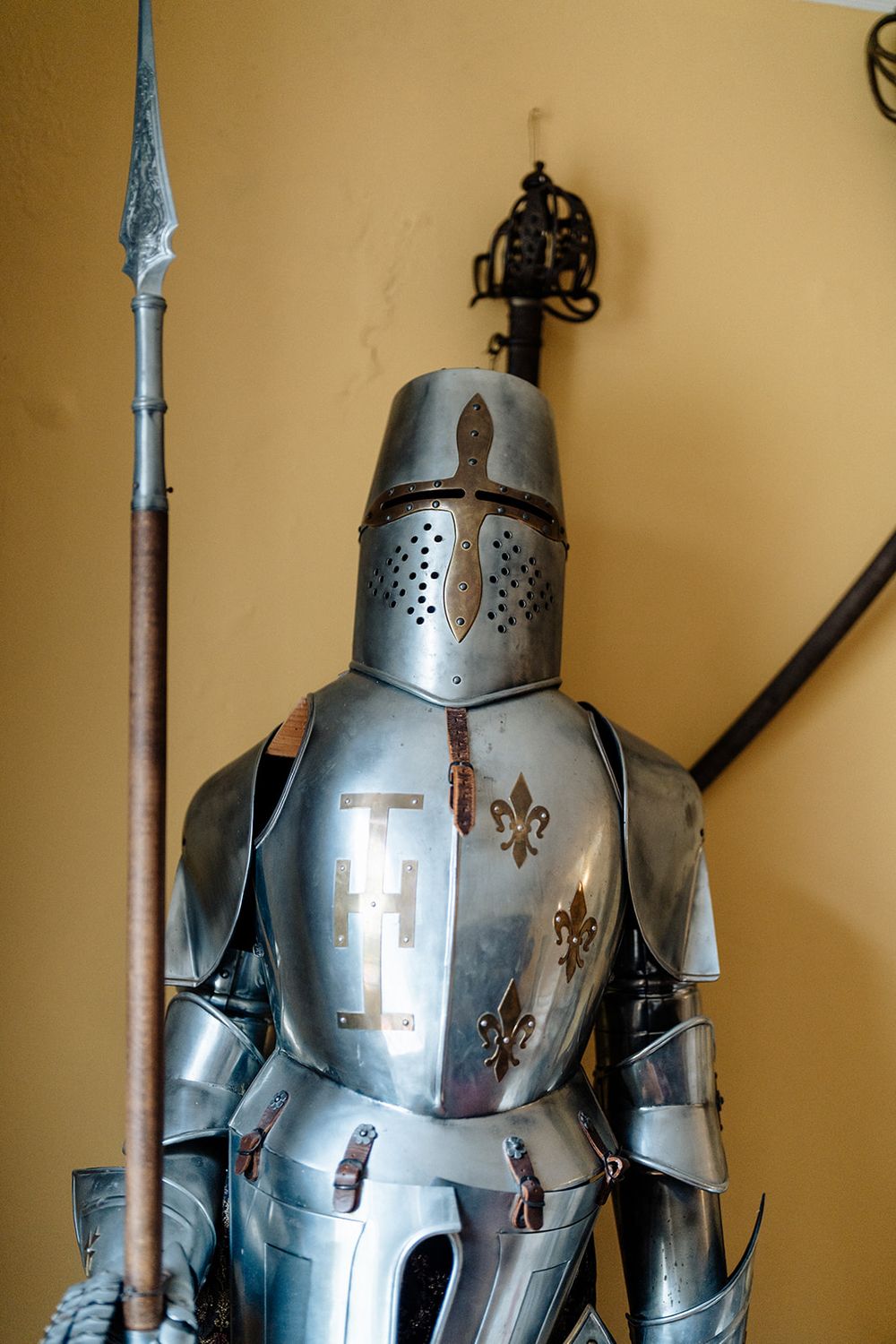 Medieval suit of armor with fleur-de-lis decorations and spear displayed against beige wall in museum setting.