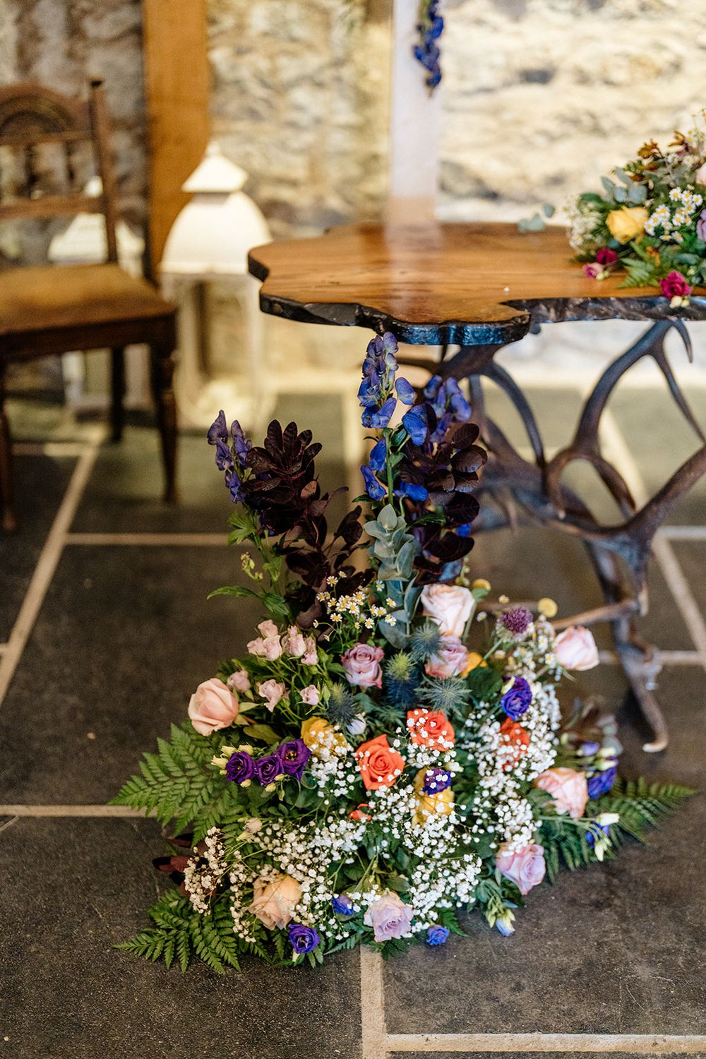Wedding floral arrangement with blue delphiniums, baby's breath, and ferns decorates a rustic wooden table.